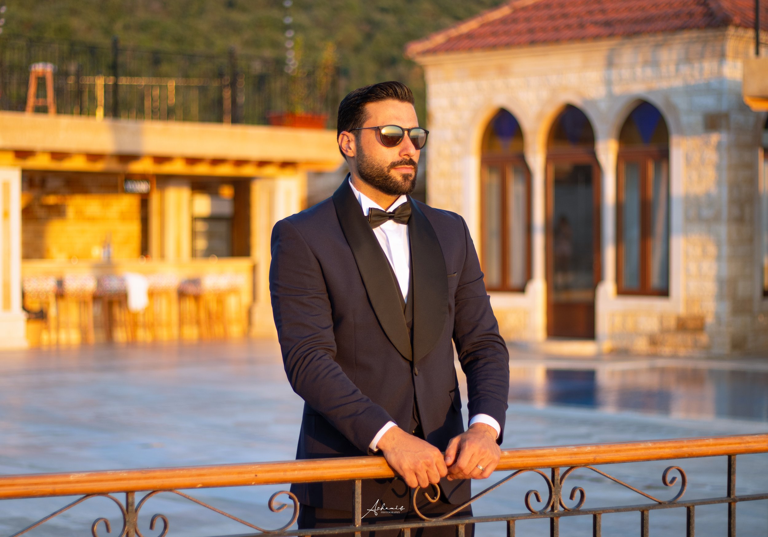 A man in a tuxedo with sunglasses standing outdoors behind a wooden railing in front of a building with stone and glass windows during sunset.