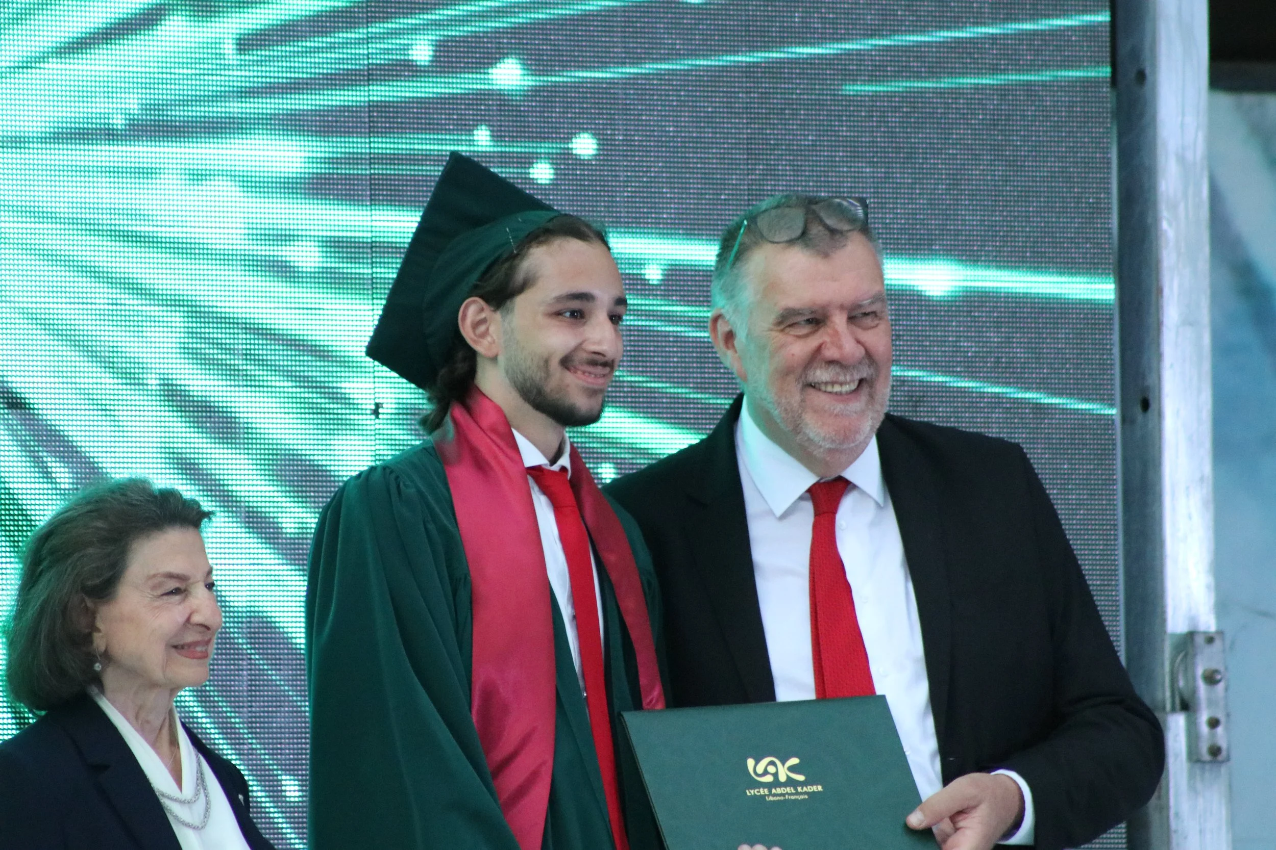A young man in a graduation gown and cap receiving a diploma from a man in a suit, with a woman also in formal attire nearby, all smiling at a graduation ceremony.