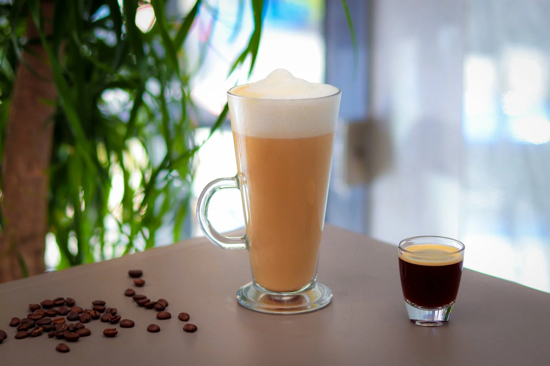 A tall glass of iced coffee with whipped cream, a small glass of espresso, and scattered coffee beans on a table.
