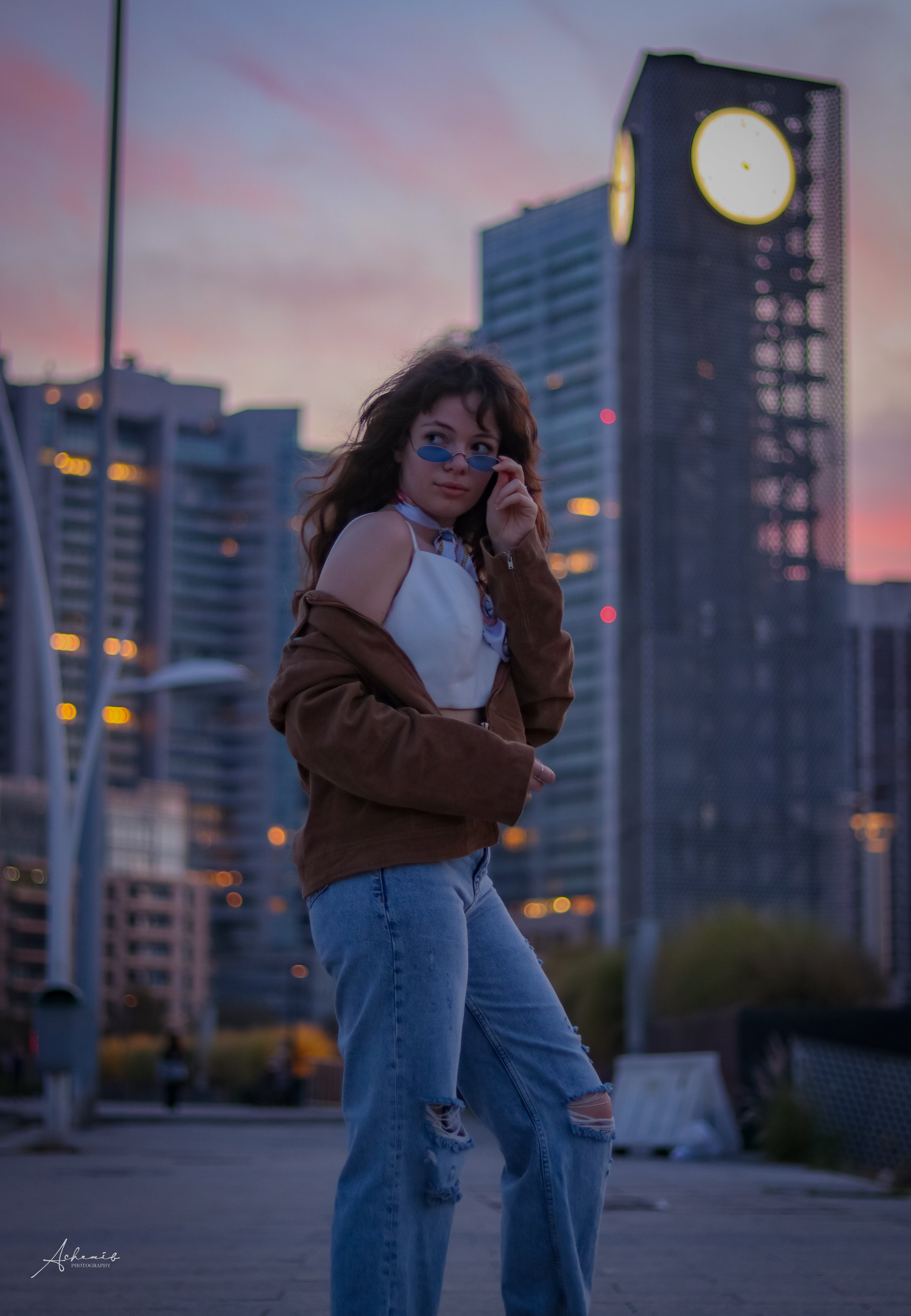 A young woman with brown wavy hair and sunglasses adjusting her glasses, wearing a white crop top, brown jacket, and ripped jeans, standing outdoors at dusk with city skyscrapers and a large clock tower in the background.