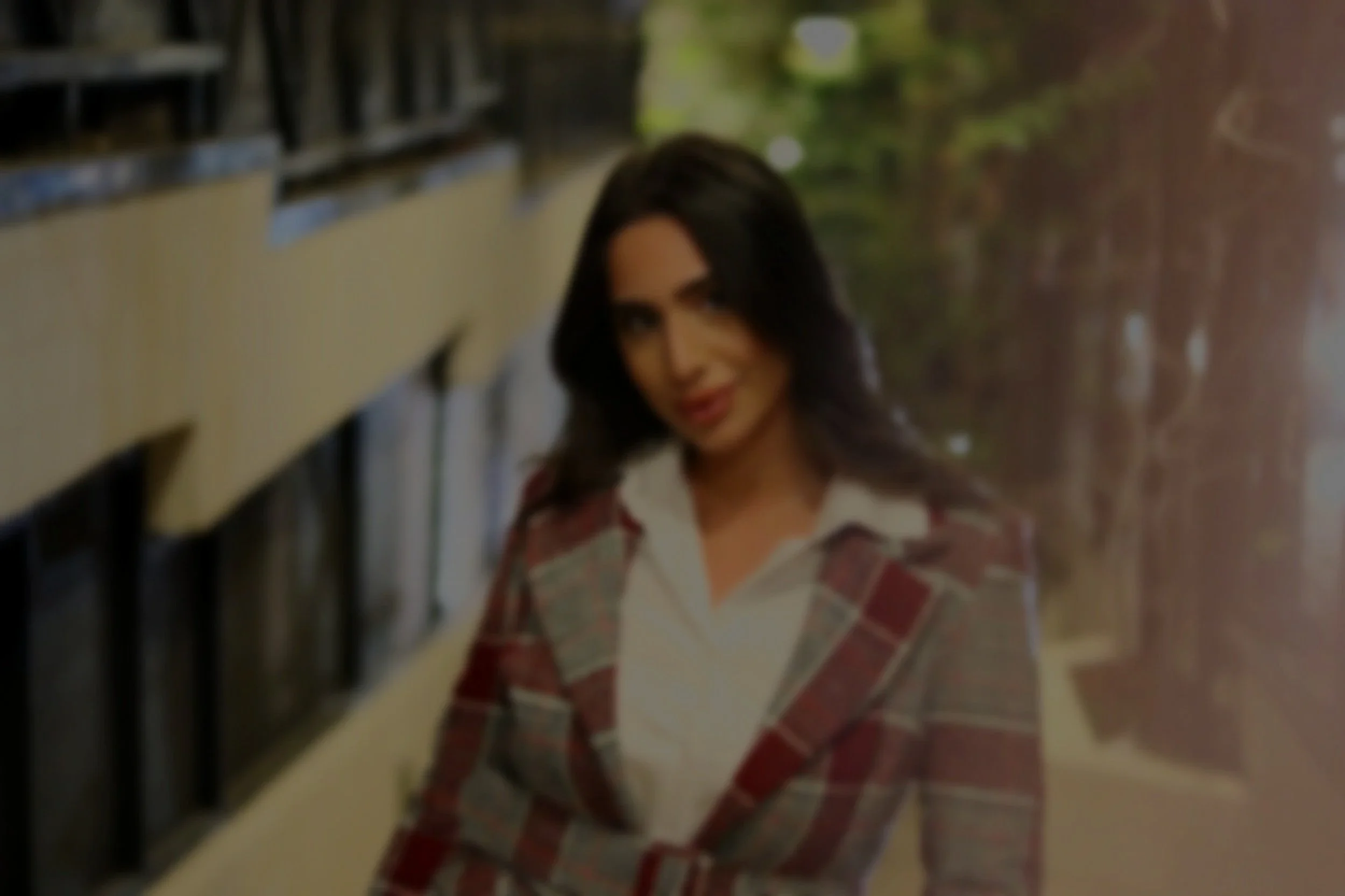 A woman with dark hair wearing a plaid blazer and white shirt, standing in front of a wall with days of the week on it, outdoors with blurred greenery in the background.