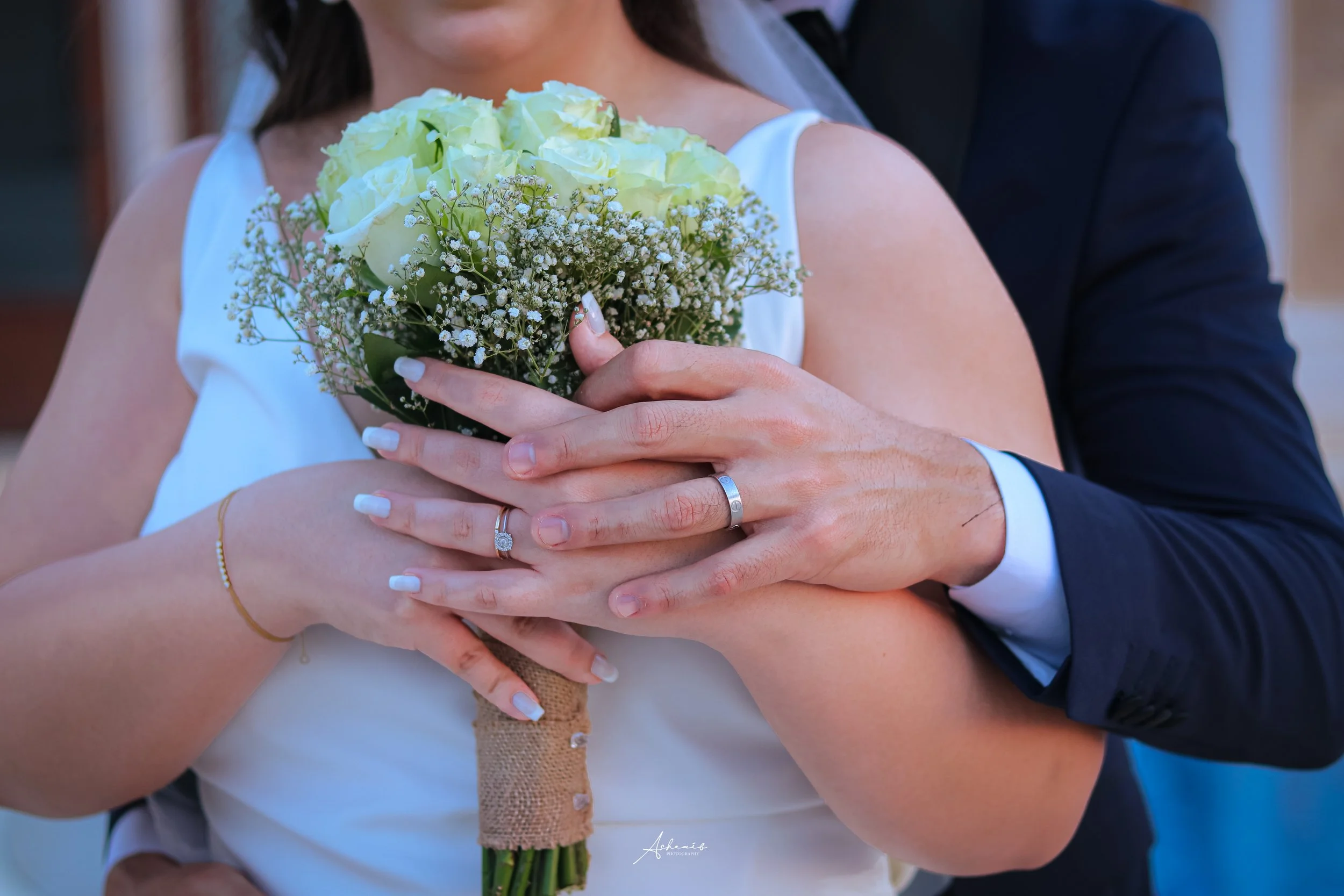 Close-up of a couple on their wedding day holding a bouquet of white roses and baby's breath, with their hands intertwined showing wedding bands.