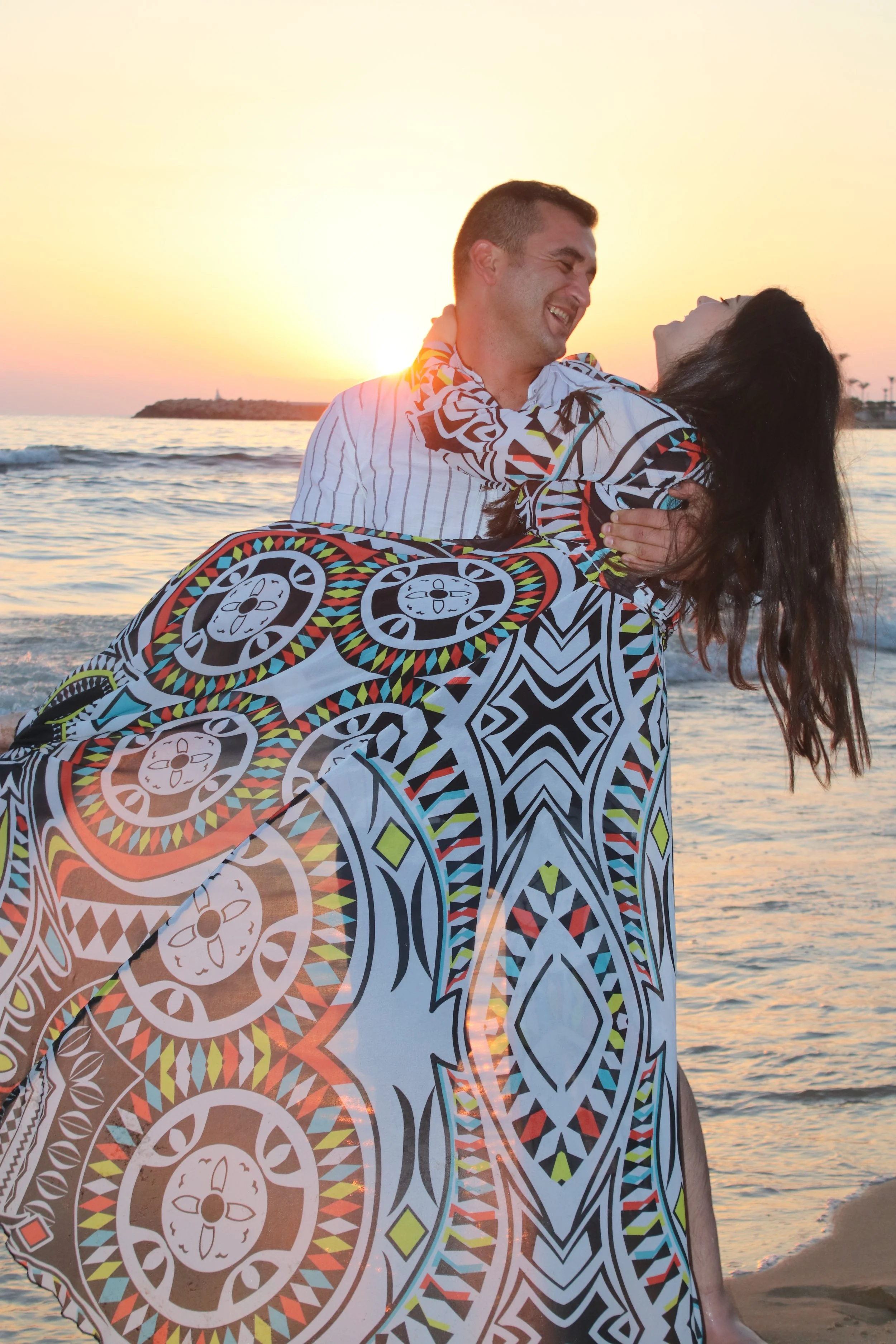 A happy couple on the beach at sunset, with the man lifting the woman in his arms. The woman is wearing a colorful patterned garment, and they are smiling at each other with the ocean and a sunset in the background.