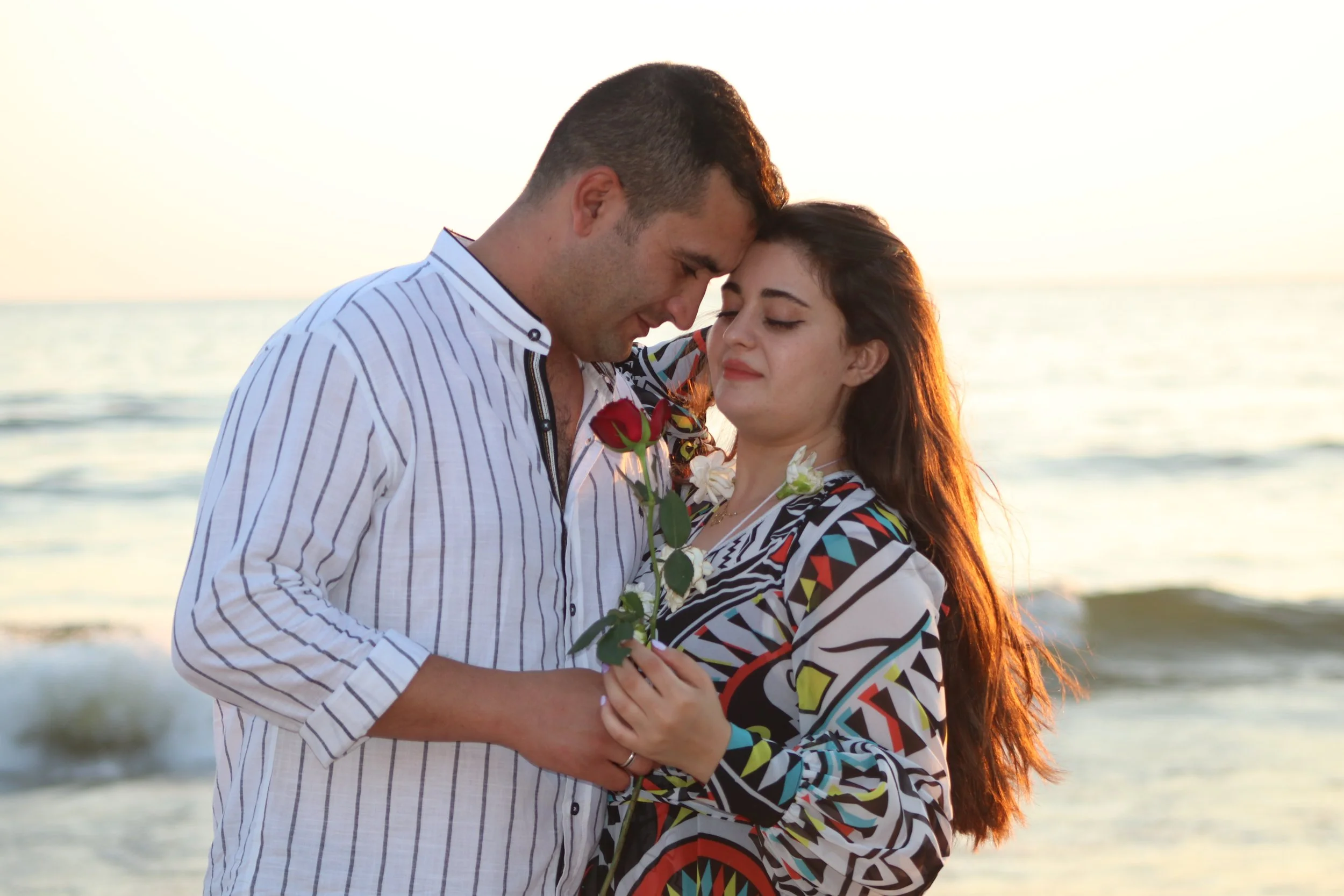A young couple stands on the beach at sunset, holding flowers and leaning their foreheads together with eyes closed, showing affection.