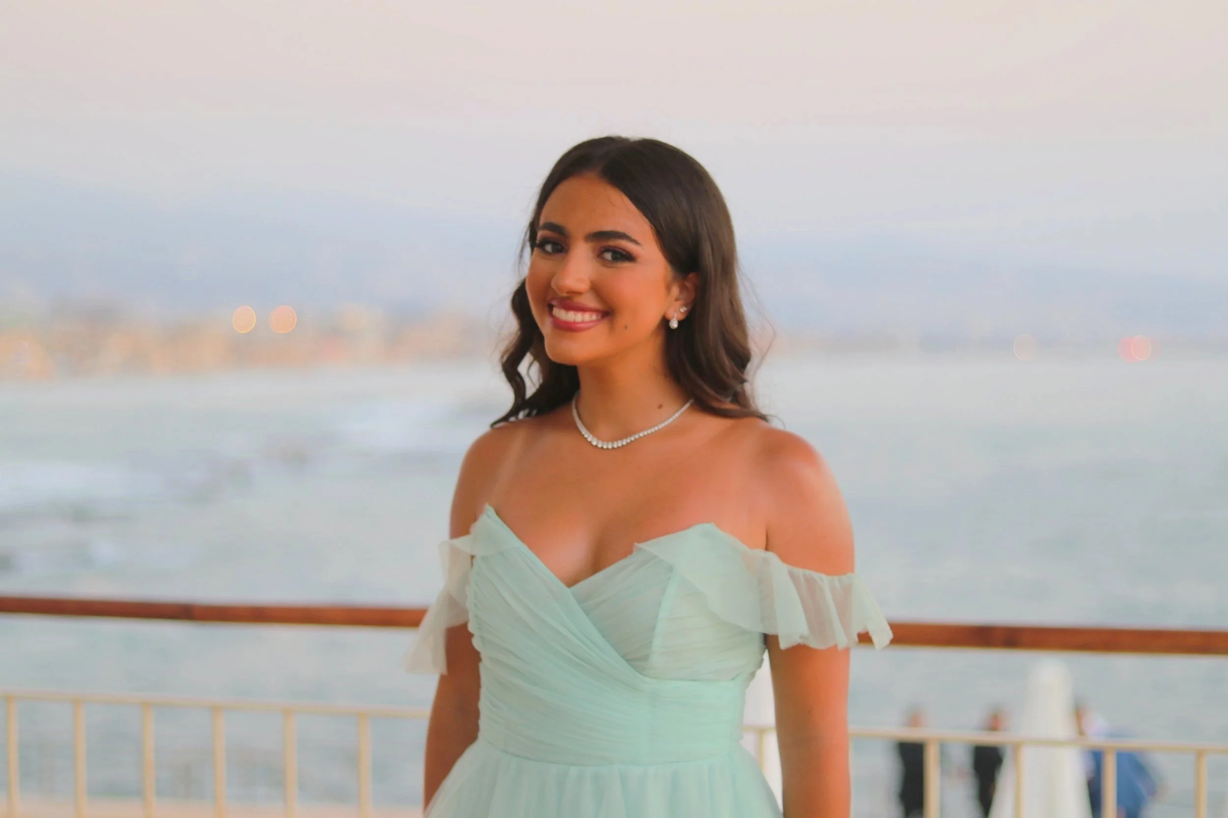 A young woman in a white off-shoulder dress standing near a railing by the ocean, smiling at the camera with a cityscape in the background during dusk.