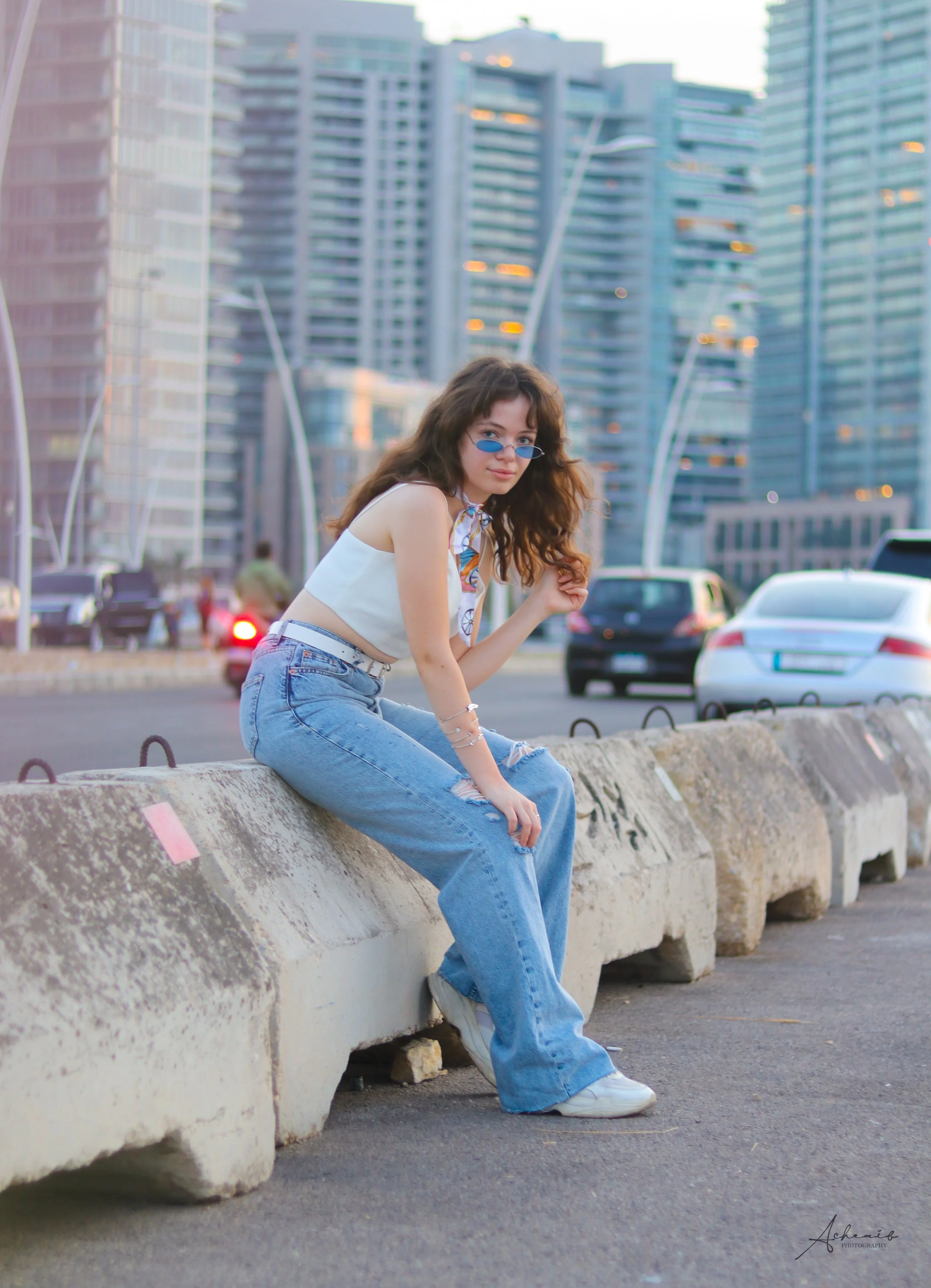A young woman with long curly hair wearing blue jeans, a white sleeveless top, and small blue sunglasses sits on a concrete barrier in an urban setting with modern skyscrapers and cars in the background.