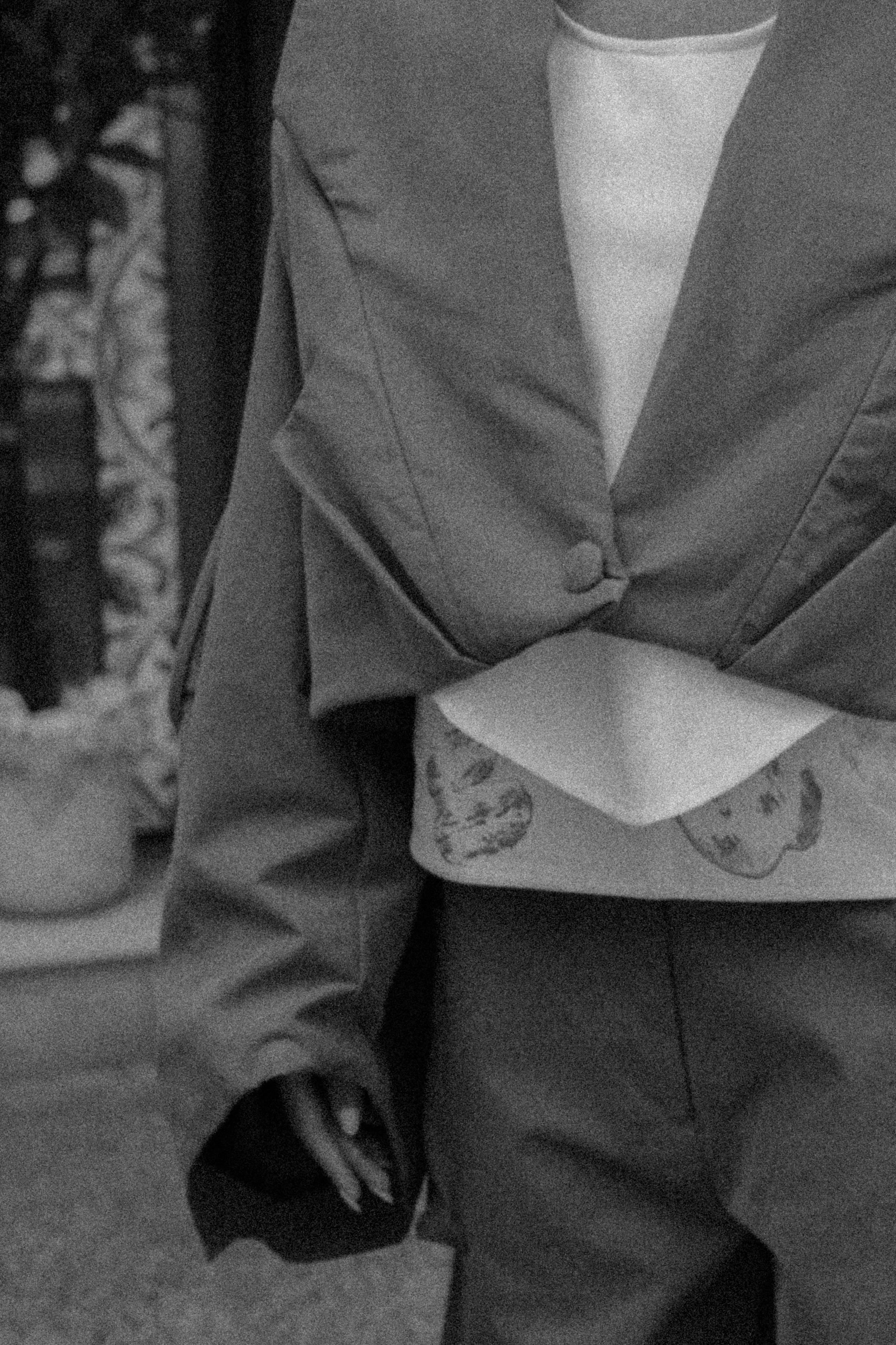 A person wearing a suit and dress shirt holding a small clutch purse, with a decorated background and a potted plant visible on the left side.