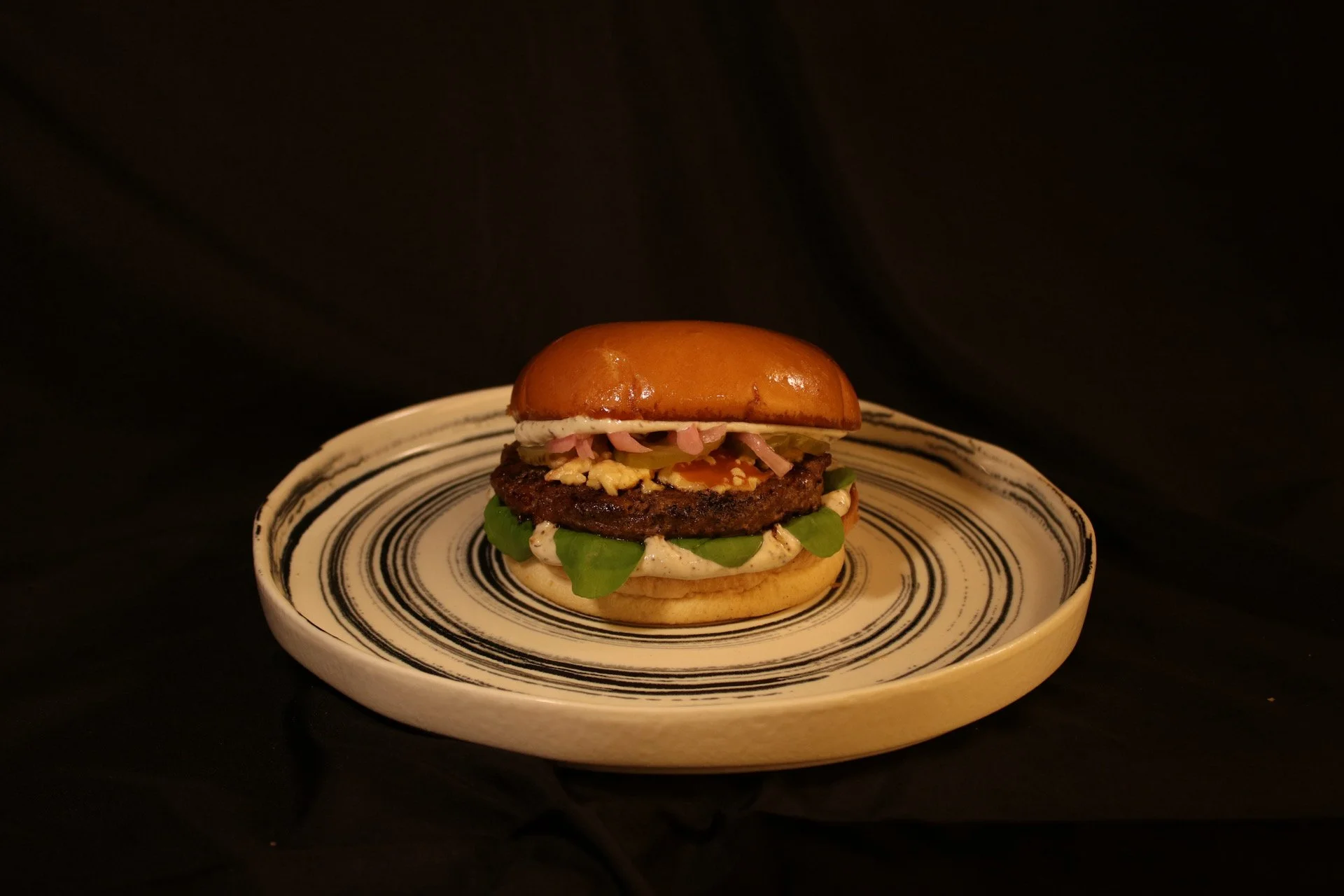 A burger with a shiny bun, lettuce, a beef patty, sliced pickles, shredded ham, and a fried egg, served on a decorative plate with black and white swirl patterns against a black background.