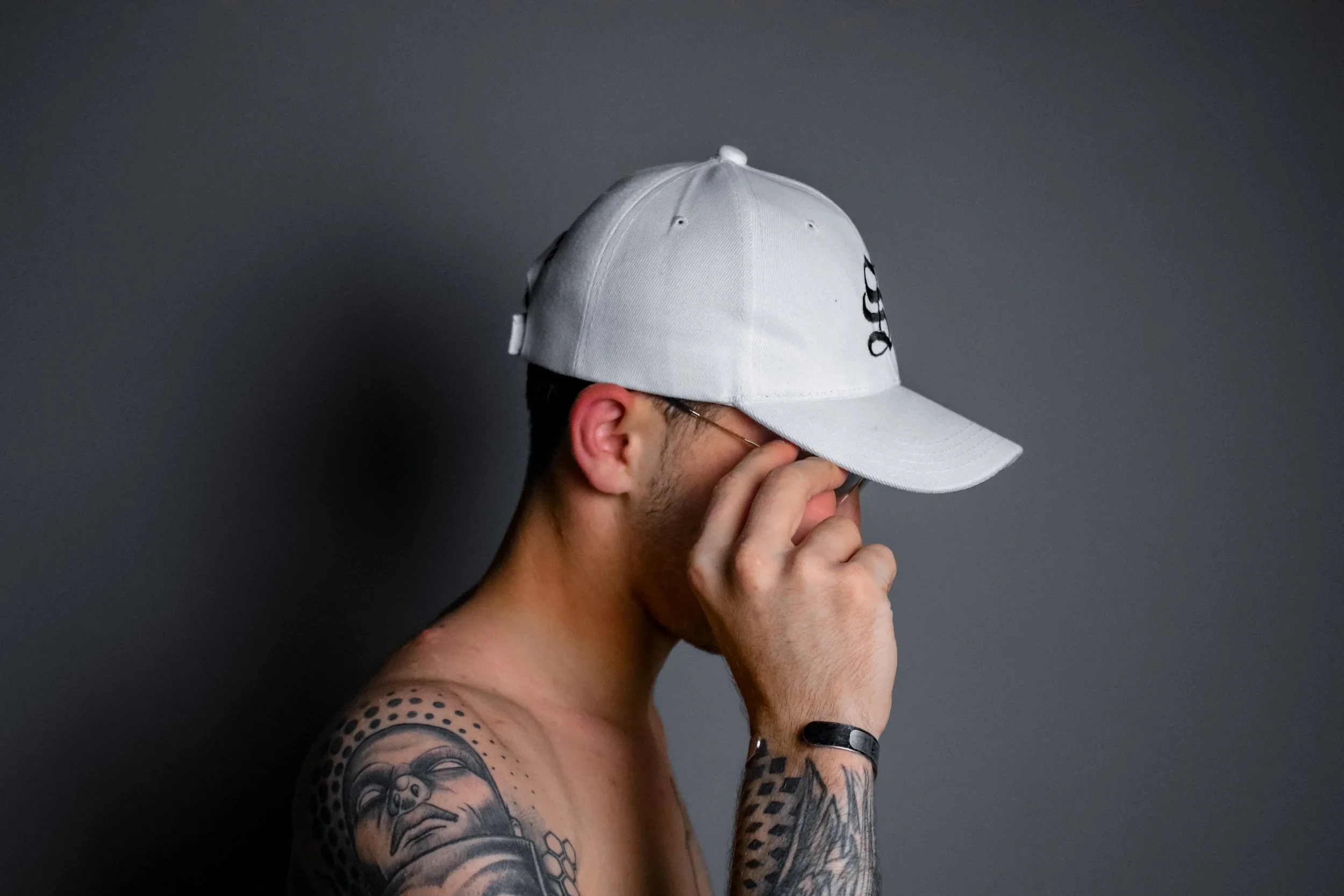 A shirtless man wearing a white baseball cap and sunglasses, with tattoos on his left arm, standing against a dark gray background.