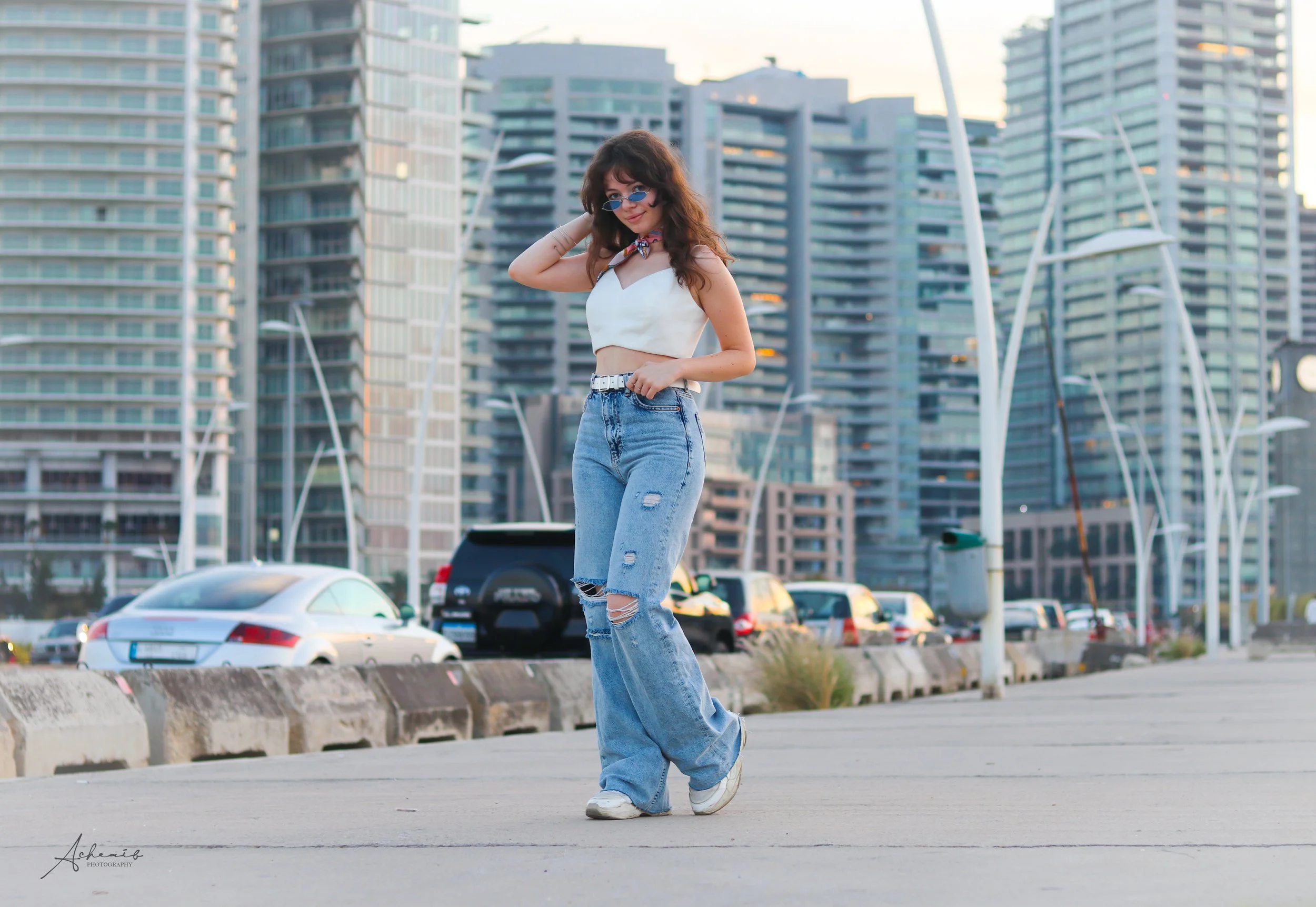 A young woman with curly brown hair wearing blue sunglasses, a white crop top, and ripped blue jeans standing on a sidewalk in an urban area with tall buildings and parked cars in the background.