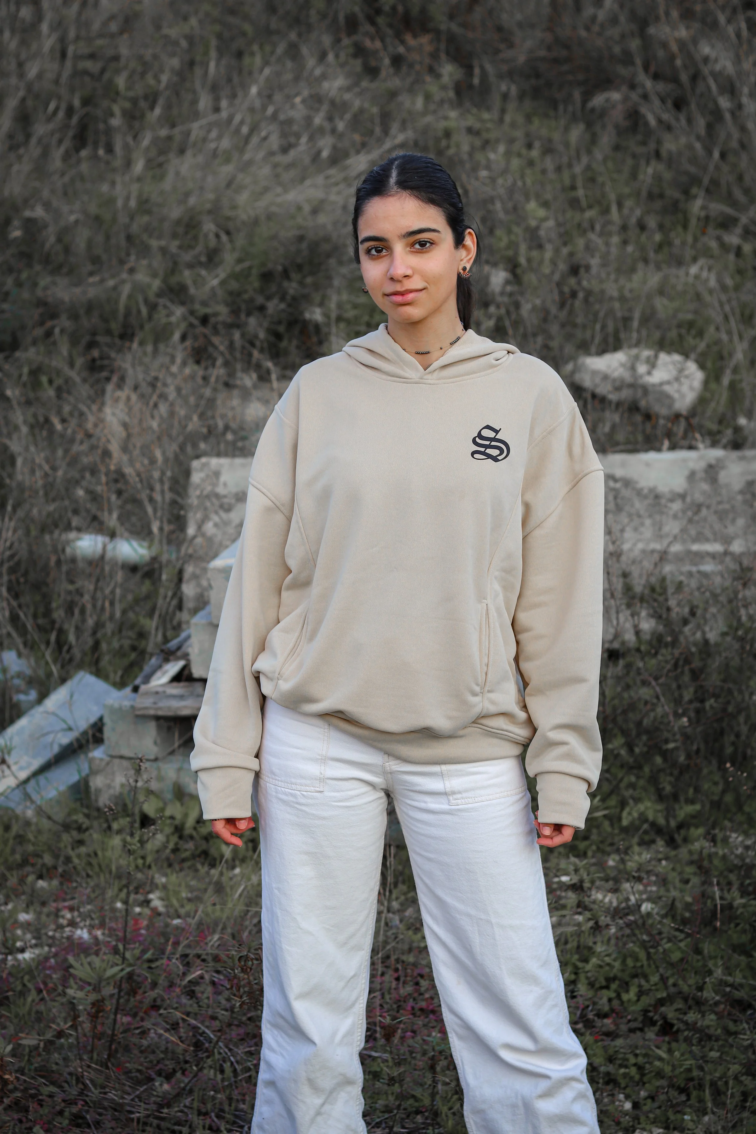 A young woman with dark hair, wearing a beige hoodie with a black print on the chest and white pants, stands outdoors against a background of dry bushes and concrete debris.