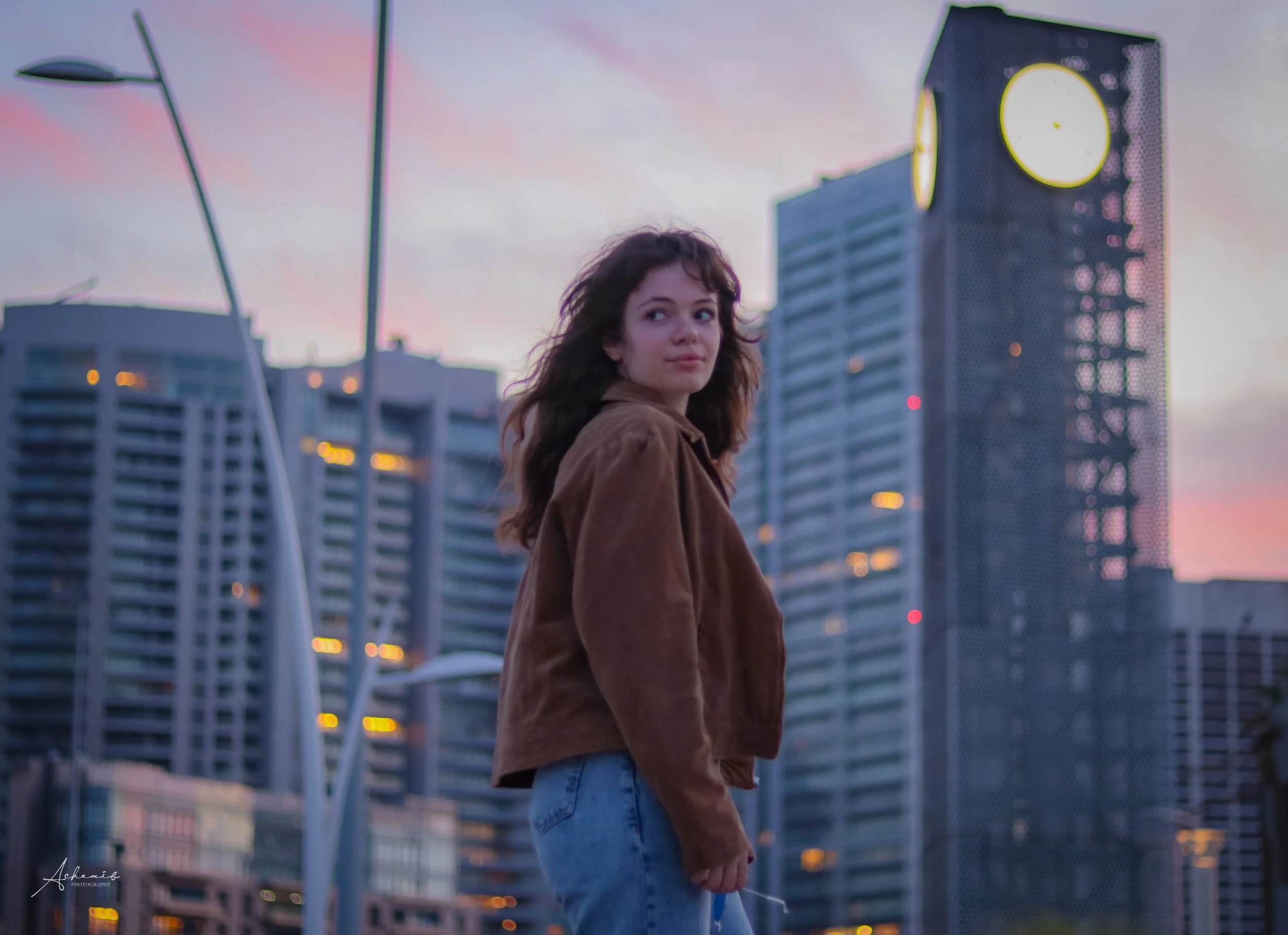 A young woman standing on an urban street during sunset, with modern high-rise buildings in the background.