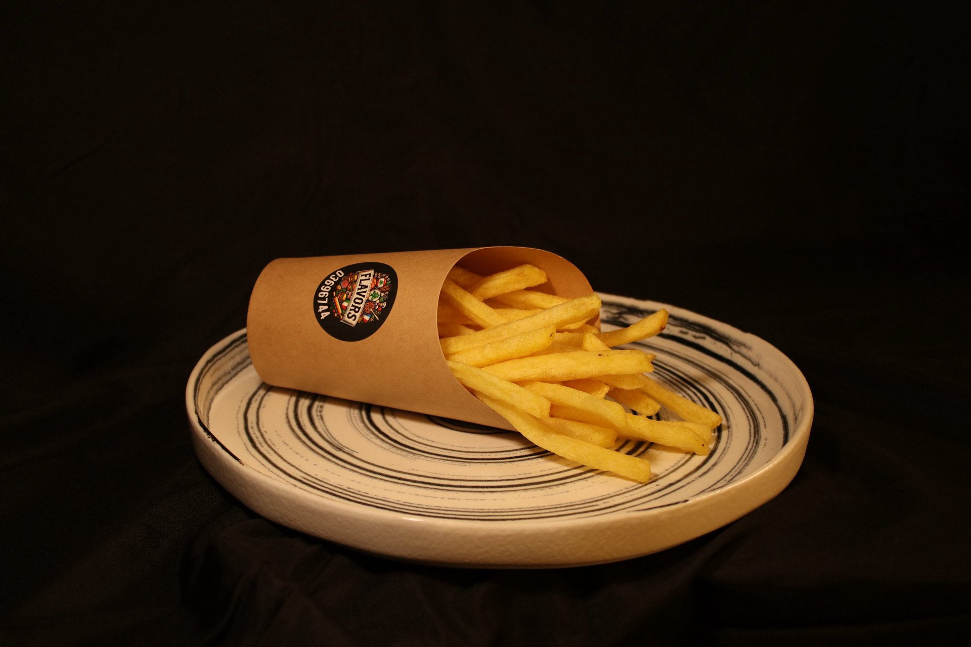 French fries in a brown paper container on a ceramic plate with black swirl pattern, against a black background.