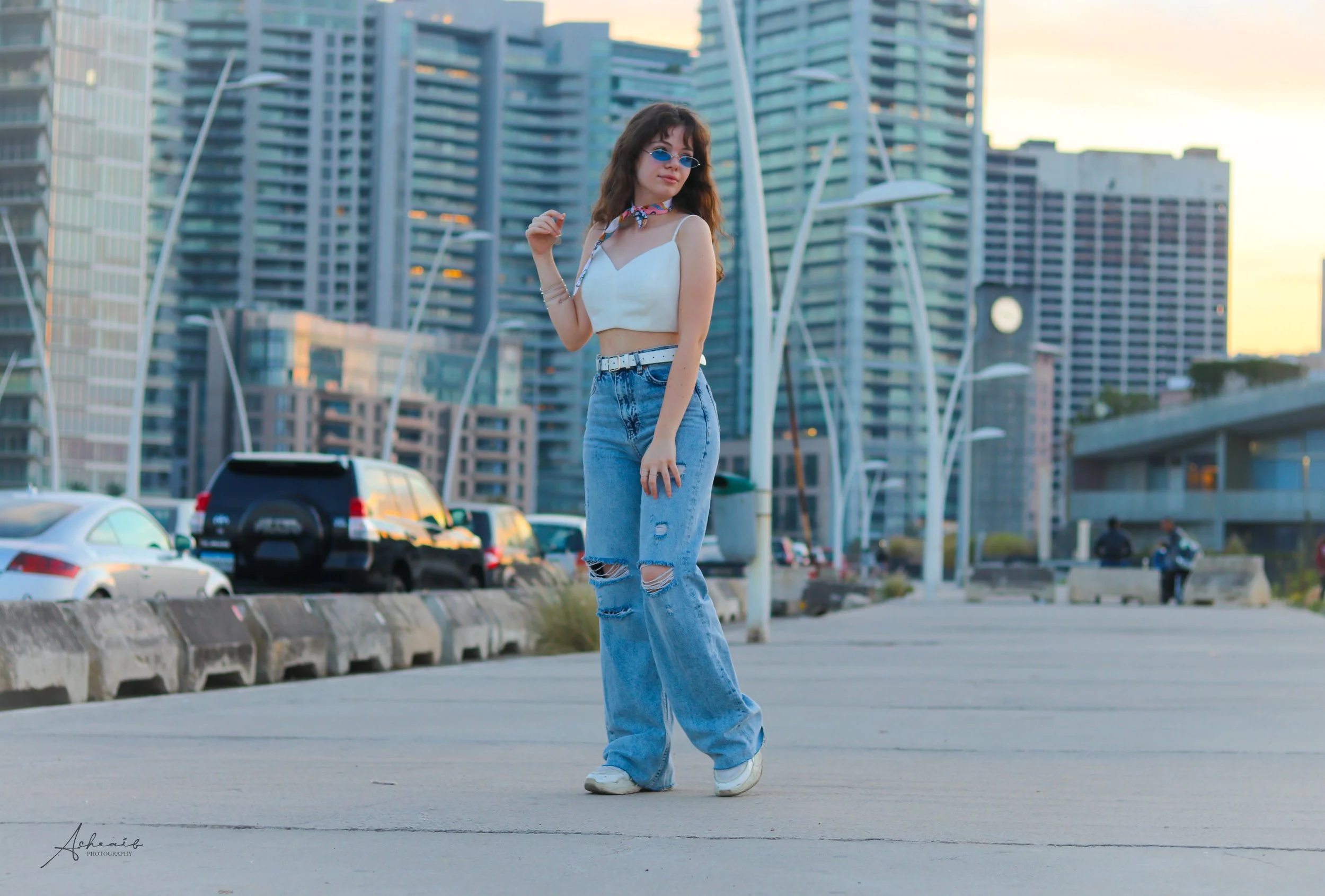 A young woman in a white crop top, ripped jeans, and sunglasses standing on a city street with modern high-rise buildings and parked cars in the background during sunset.