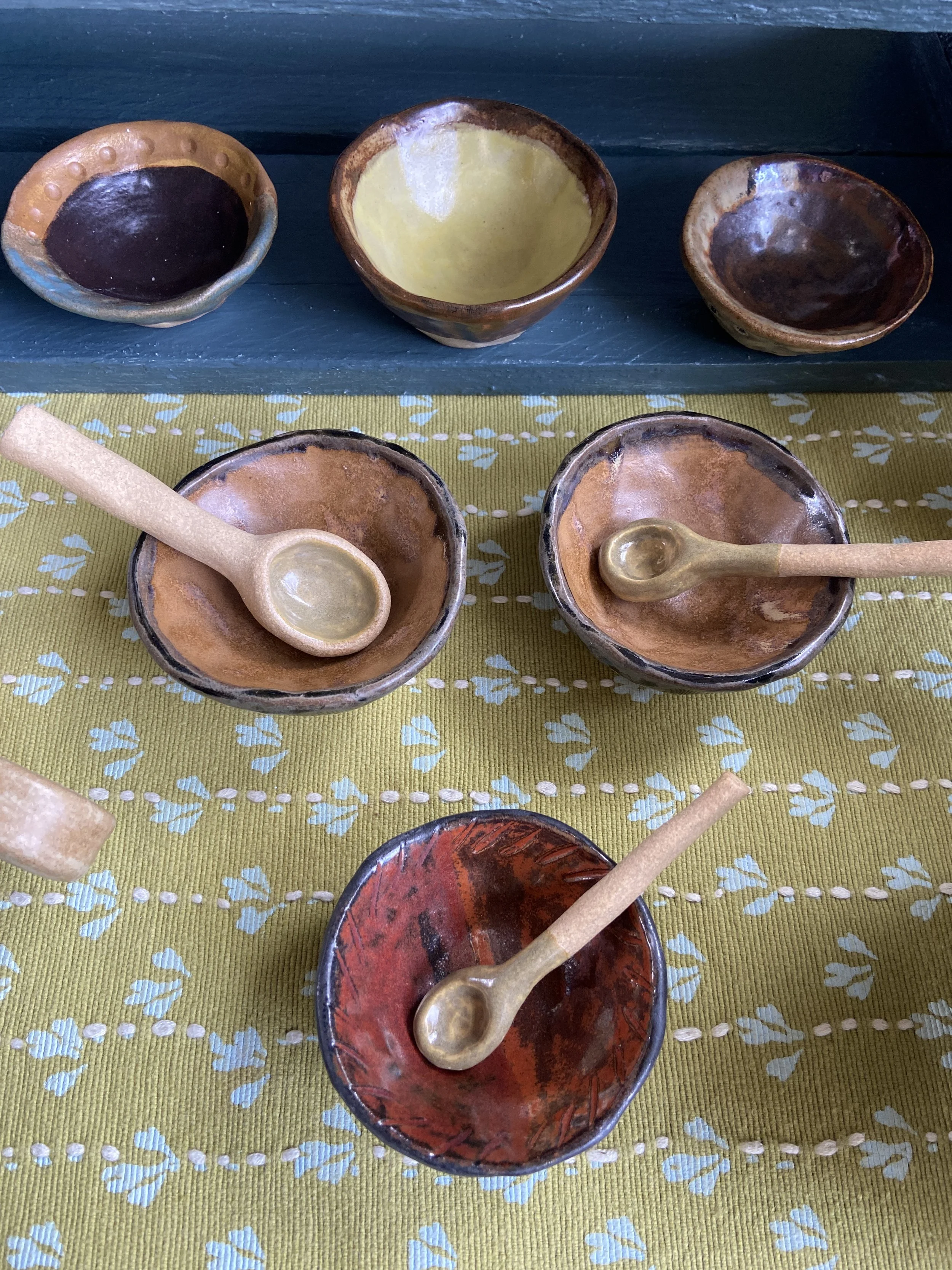 Six small, colorful ceramic bowls arranged on a table with three of them containing light-colored ceramic spoons. The bowls have various designs and are positioned on a green tablecloth with a white and yellow floral pattern. A dark blue wooden shelf or board with three additional bowls is in the background.