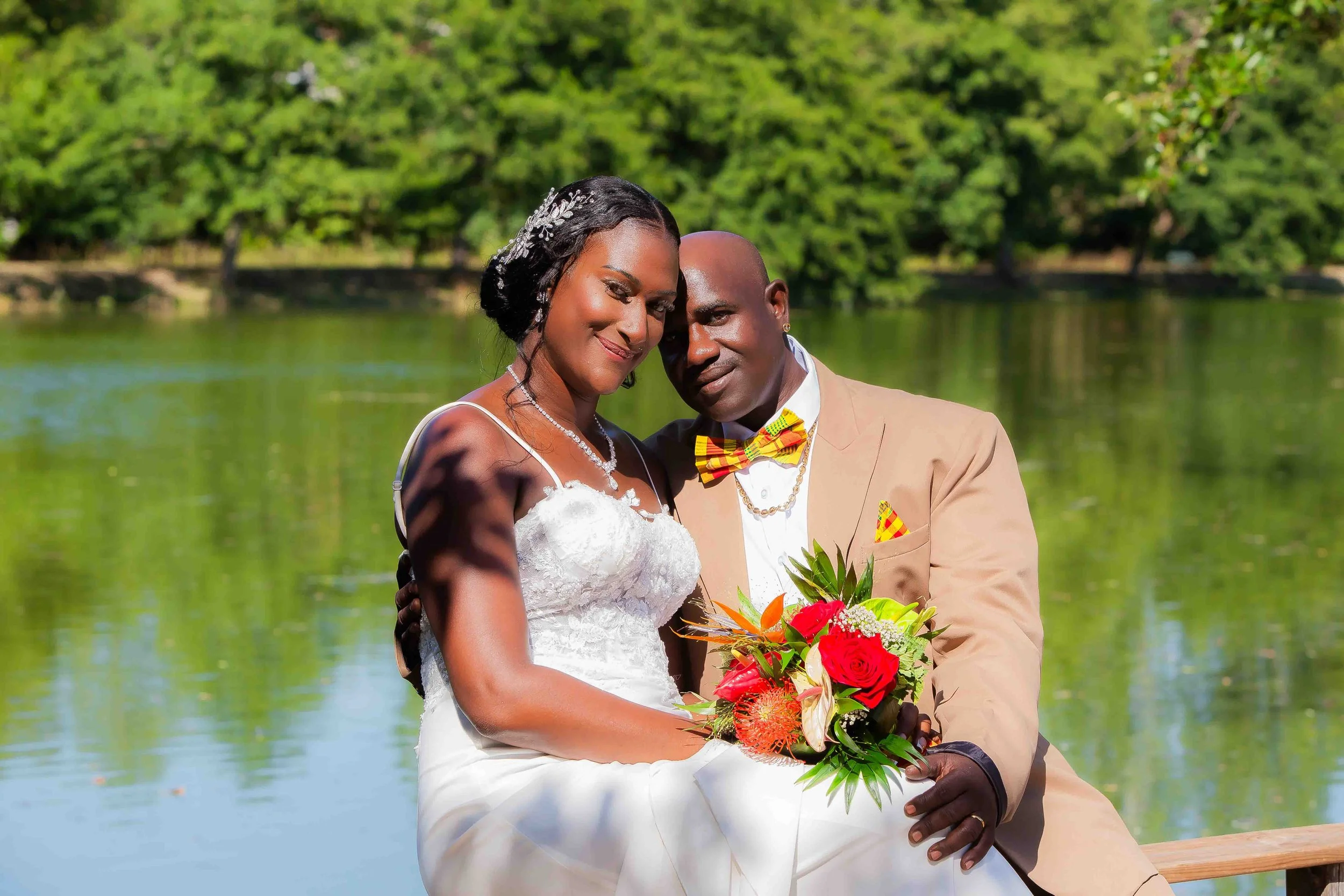 Un couple se tenant par la main lors d'un mariage en plein air, avec un lac et des arbres verts en arrière-plan, la femme en robe de mariée blanche et l'homme en costume beige avec un nœud papillon à motifs
