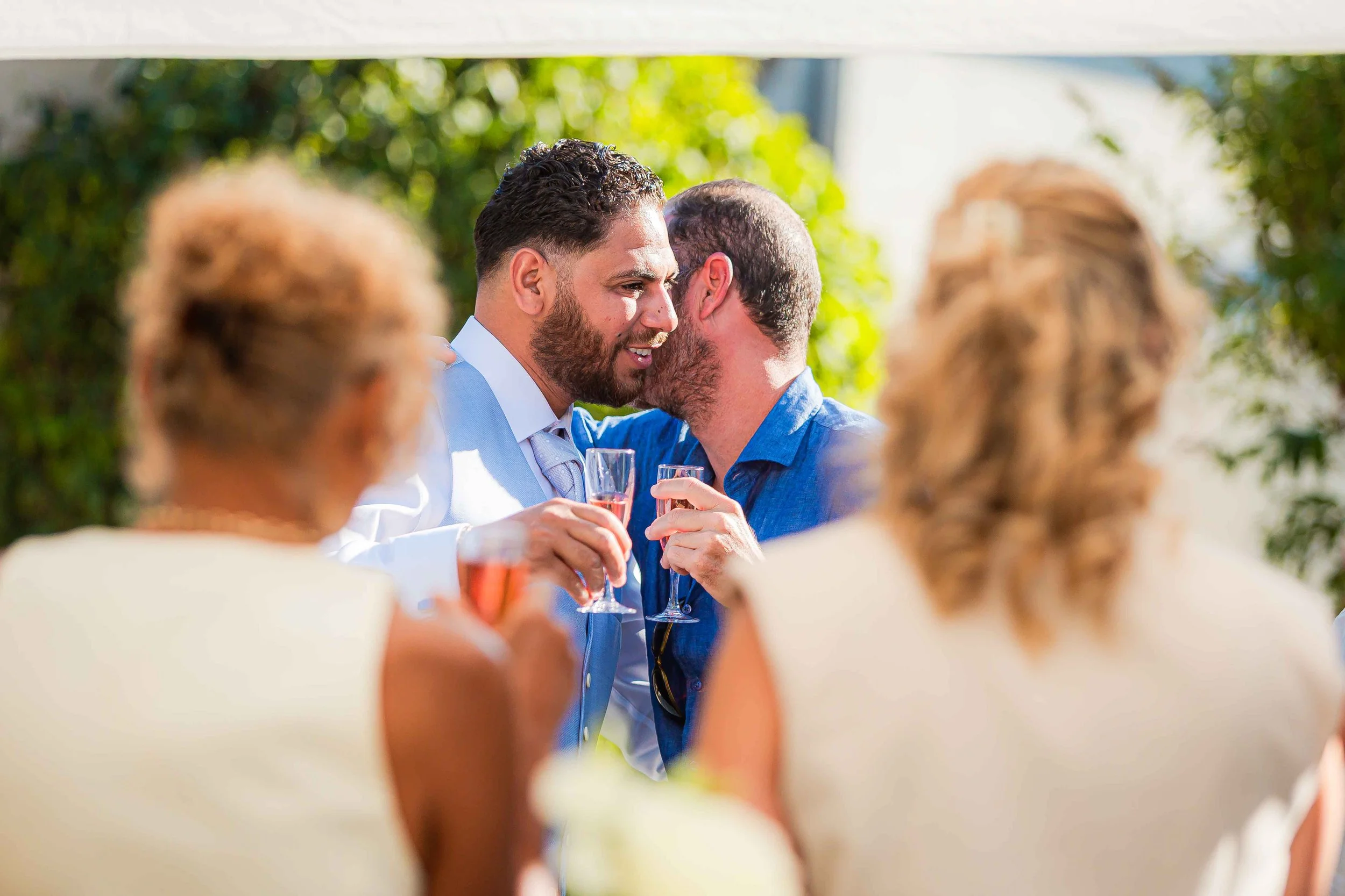 Deux hommes en costume qui échangent un toast lors d'une célébration en plein air, entourés d'invités en tenue élégante et de végétation luxuriante.