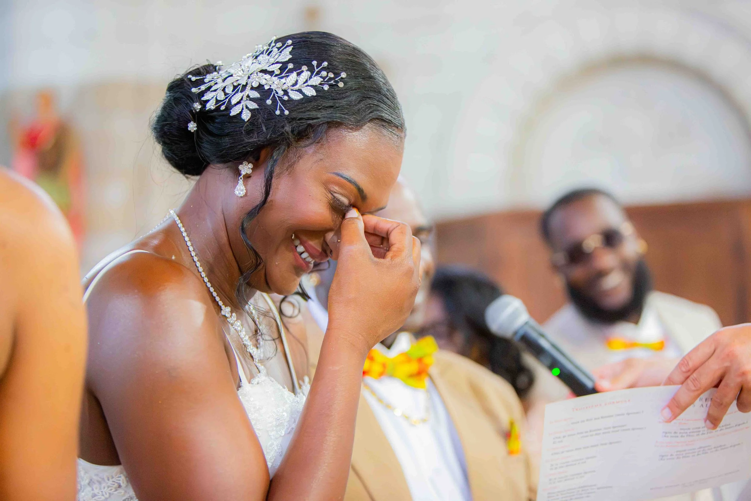 Une femme en robe de mariée, coiffée d'un chignon avec un-accessoire en argent, pleure de joie lors d'une cérémonie, entourée d'autres personnes.