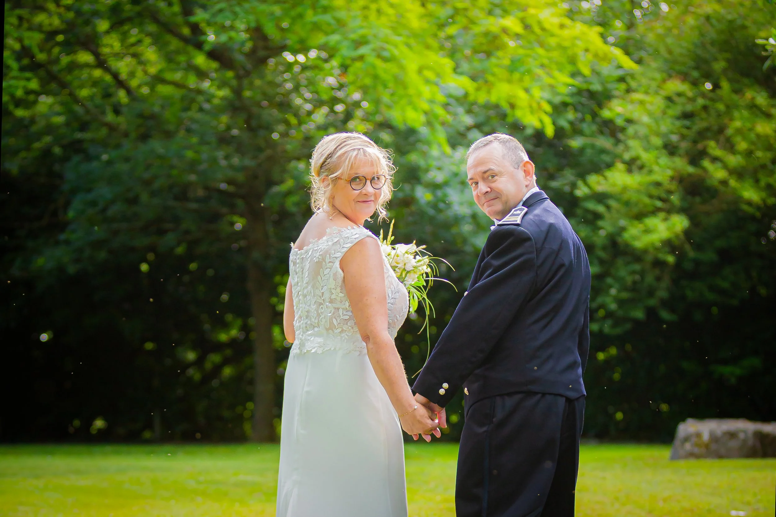 Un couple en mariage, tenant la main dans un parc verdoyant, regardent en arrière, avec des arbres en toile de fond.