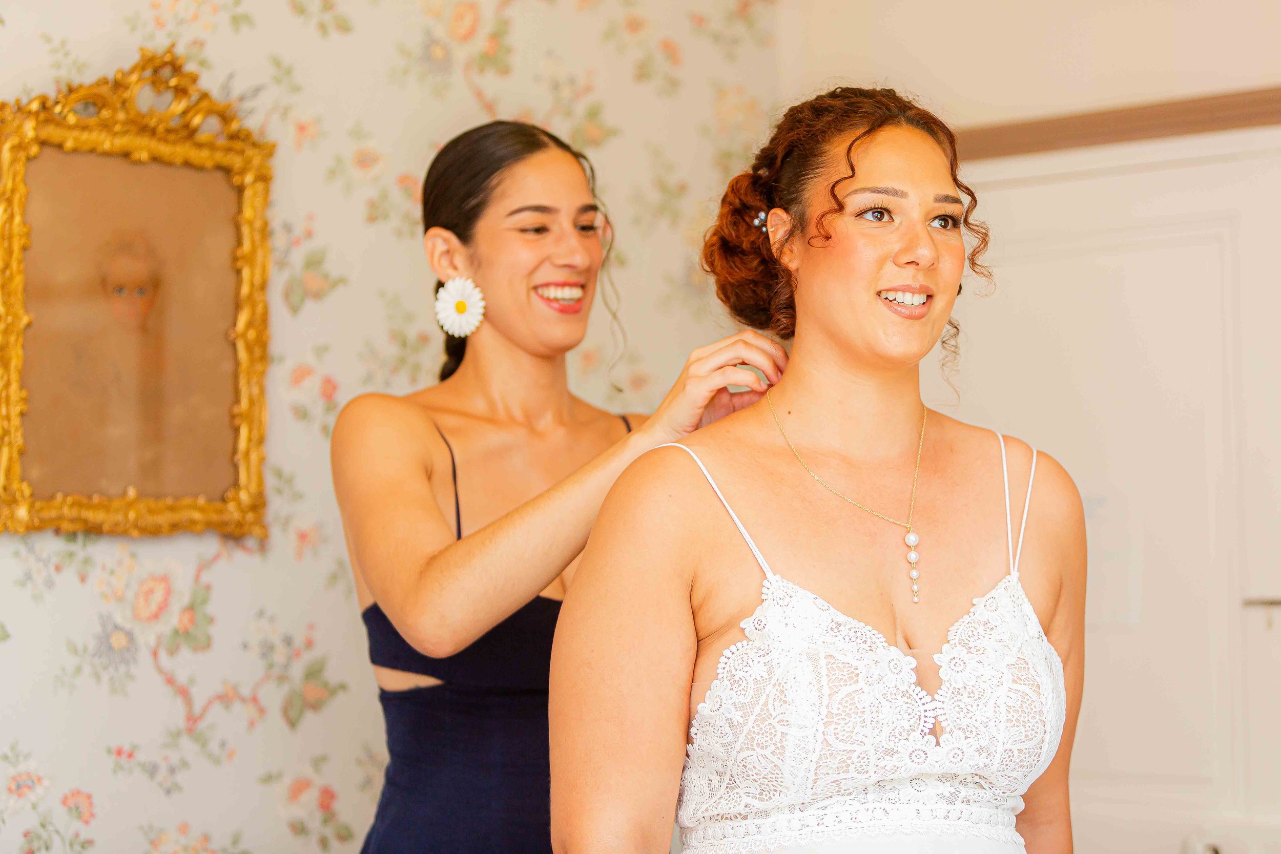 Deux femmes en train de préparer une mariée, une ajustant sa robe de mariée, dans une pièce décorée avec un mur à motif floral et un grand cadre doré.