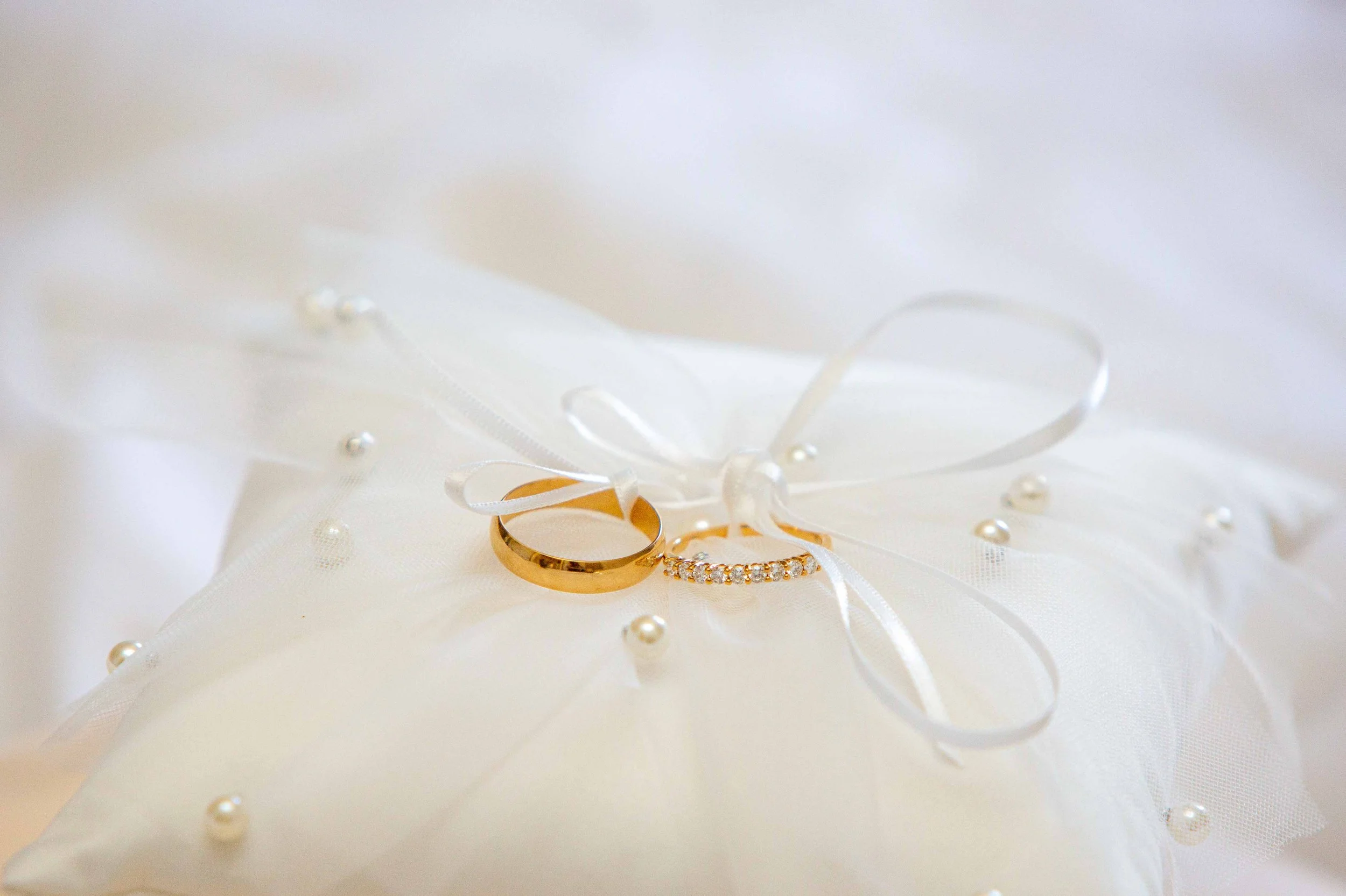 Deux alliances dorées posées sur un tissu blanc, entourées de rubans satinés et de perles. Un décor de mariage.