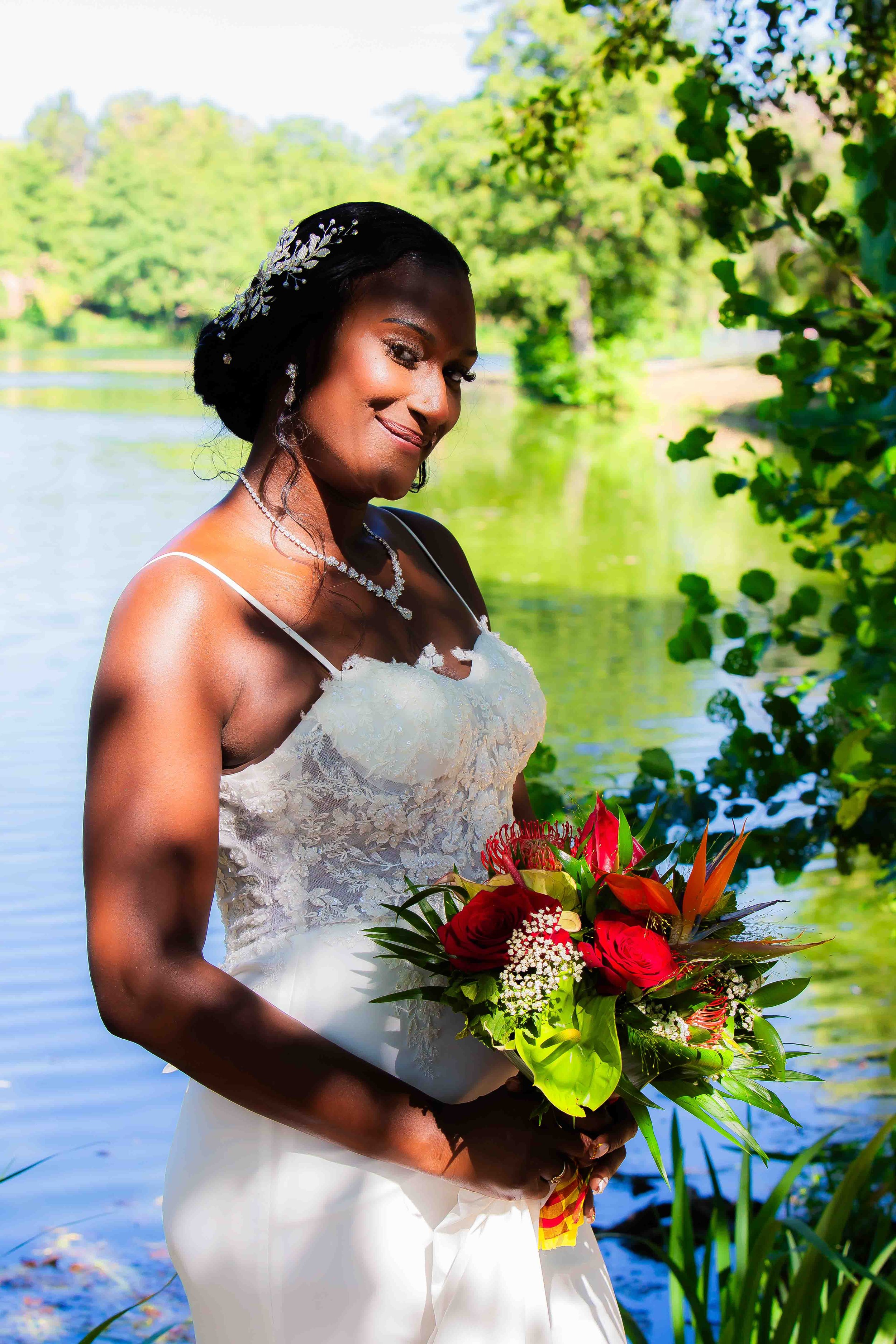 Une femme en robe de mariage blanches, portant un collier et des boucles d'oreilles, tient un bouquet de fleurs rouges et vertes devant un lac entouré d'arbres en été, sous un ciel ensoleillé.