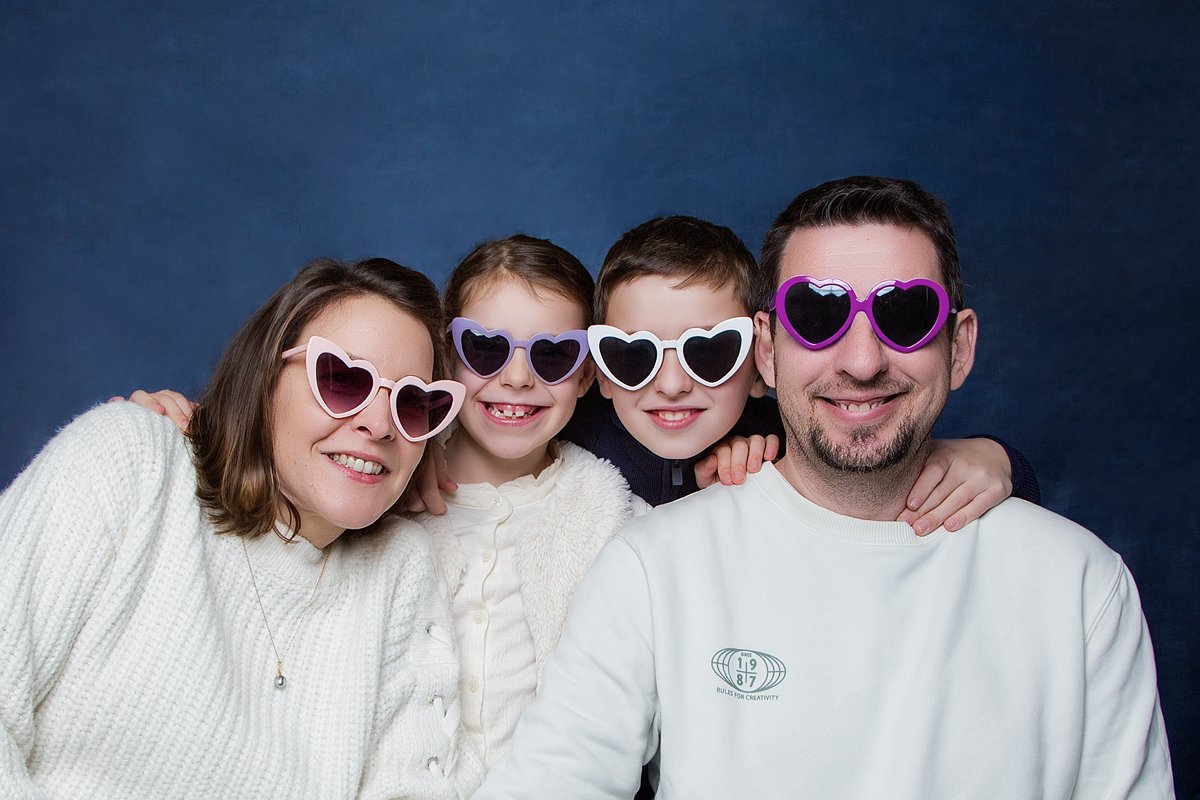 Une famille de quatre personnes porte des lunettes de soleil en forme de cœur, souriant, contre un fond bleu.