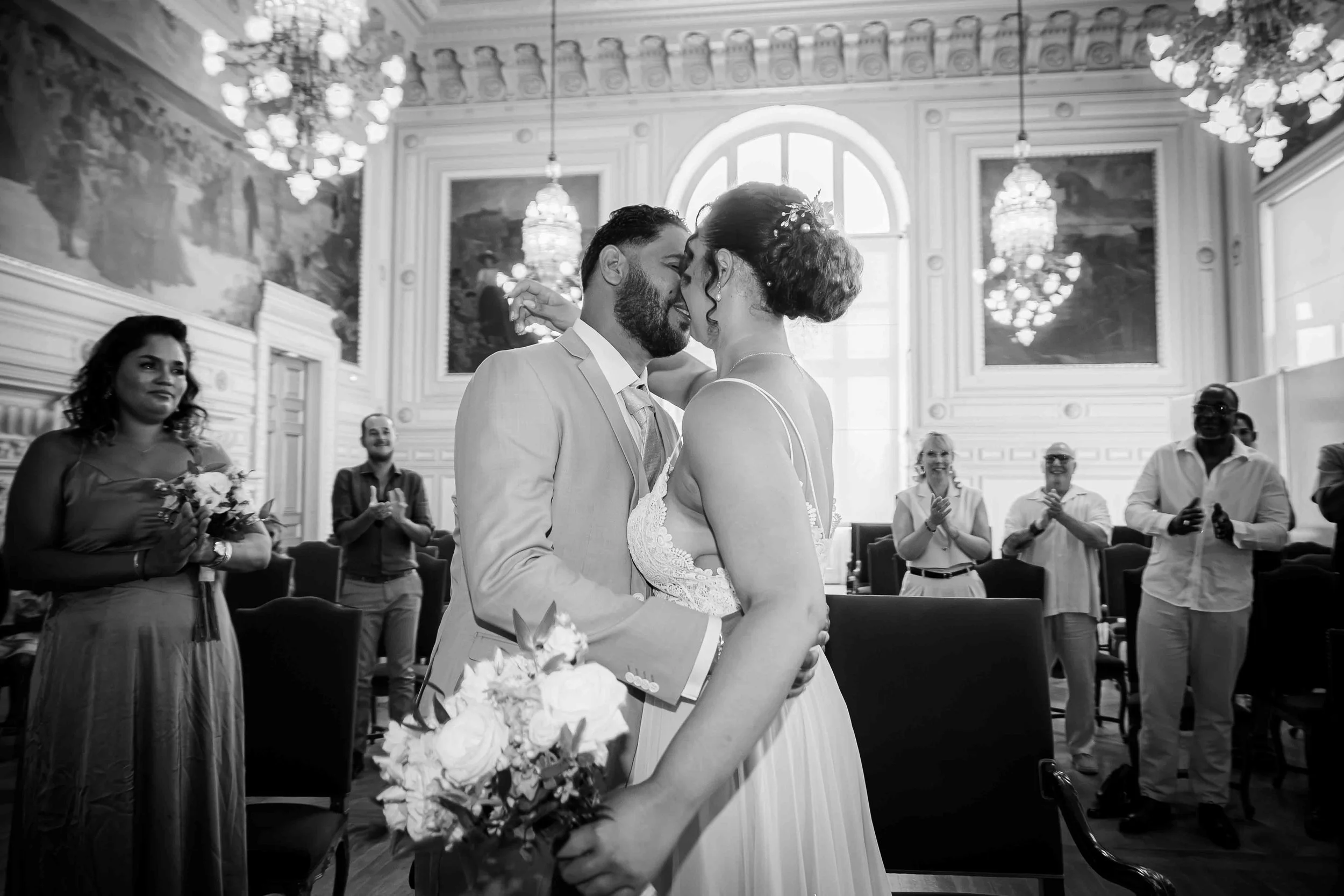 Un couple de mariés s'embrassant lors de leur mariage dans une salle élégante, entourés de témoins et amis, avec des lumières chandelier et un décor classique.