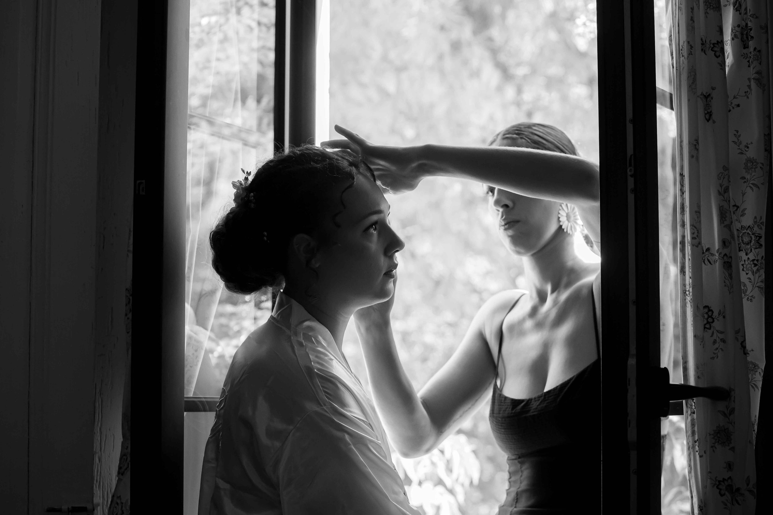 Deux femmes, dont une probablement une mariée, dans une pièce près d'une fenêtre, en train de se maquiller ou de se coiffer, dans un style noir et blanc.