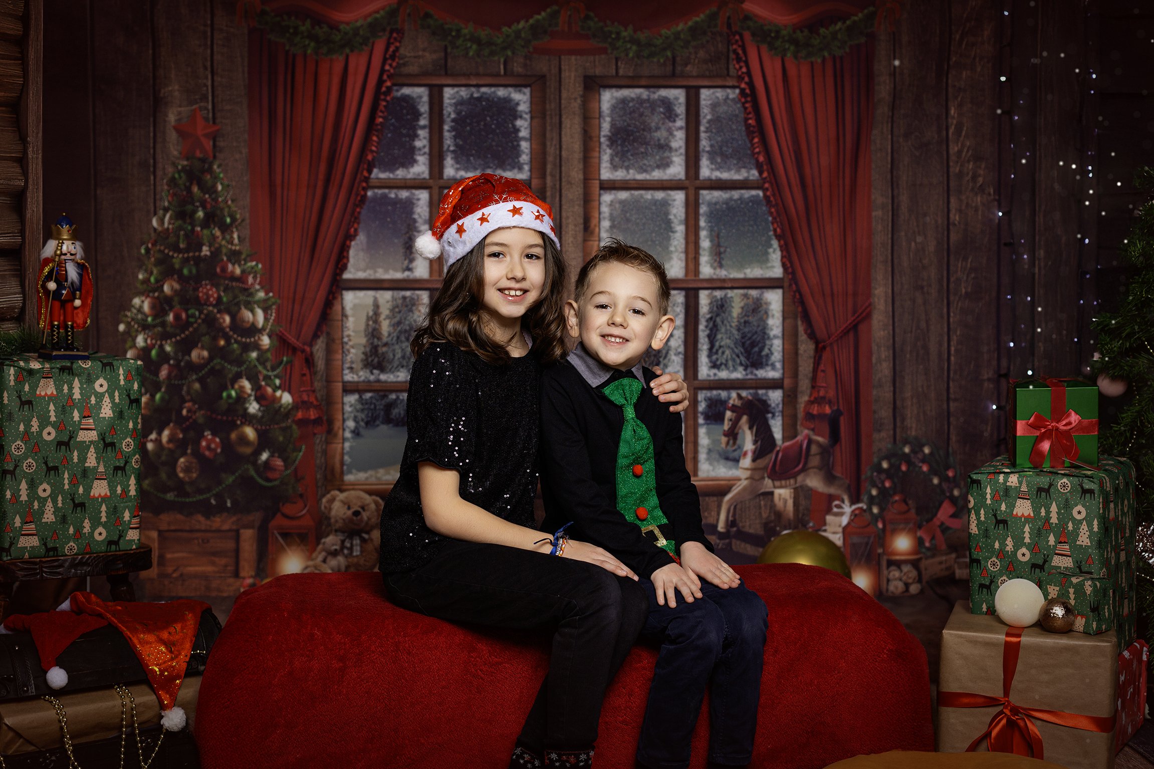 Deux enfants assis sur un lit rouge dans une scène décorée pour Noël. La fille porte un bonnet de Père Noël et une robe noire avec des paillettes, le garçon porte un pull noir avec une cravate verte et une moustache de Noël. En arrière-plan, il y a un sapin de Noël décoré, des cadeaux, une fenêtre avec un paysage hivernal enneigé, des guirlandes lumineuses et des décorations festives.