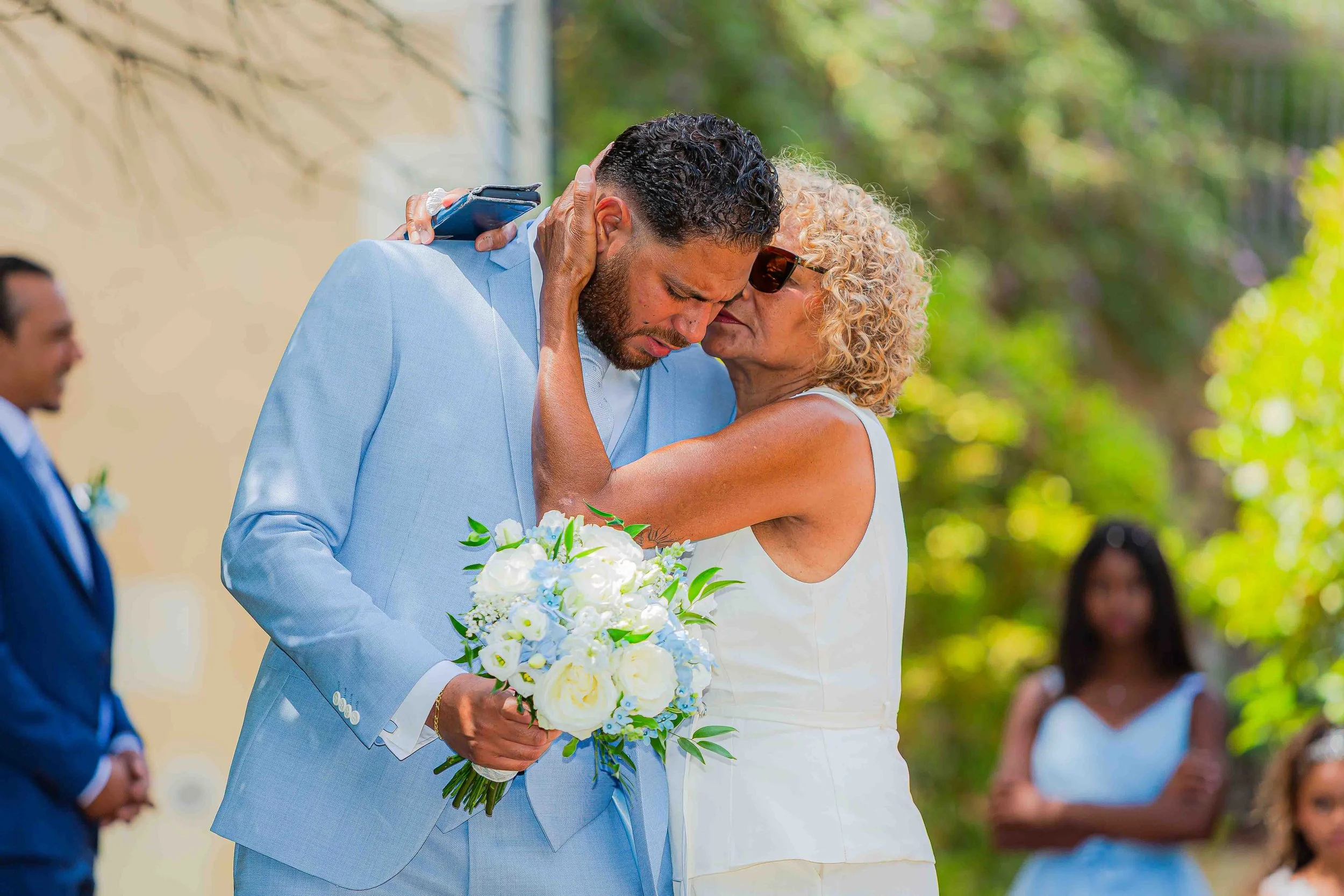 Une femme âgée avec des cheveux blonds frisés et portant des lunettes embrasse un homme avec des cheveux noirs bouclés et une barbe lors d'une cérémonie de mariage en plein air. Il porte un costume bleu clair et elle tient un bouquet de fleurs blanch