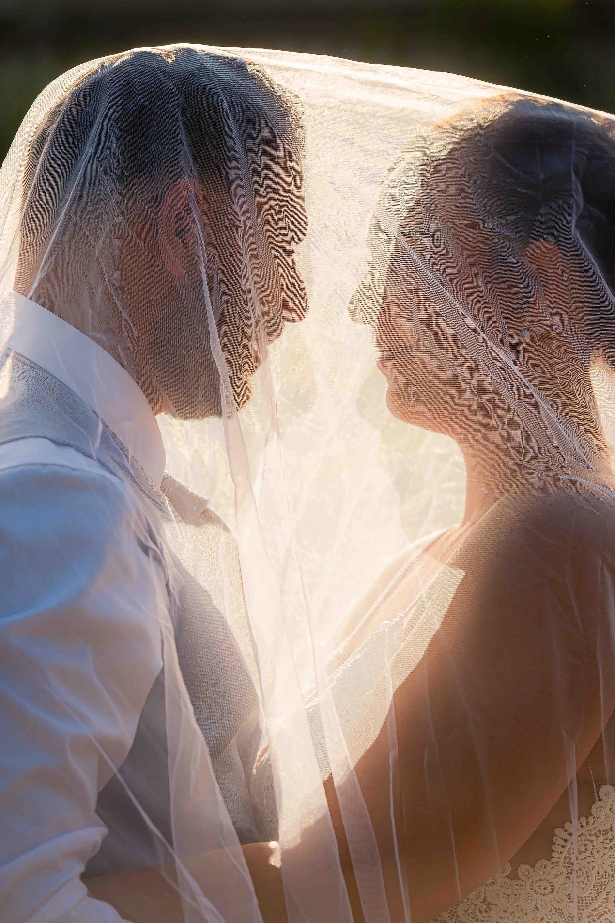 Un couple intime devant la lumière du soleil, la femme porte une robe blanche avec dentelle et le voile de la mariée, l'homme porte une chemise blanche. Ils se regardent tendrement.