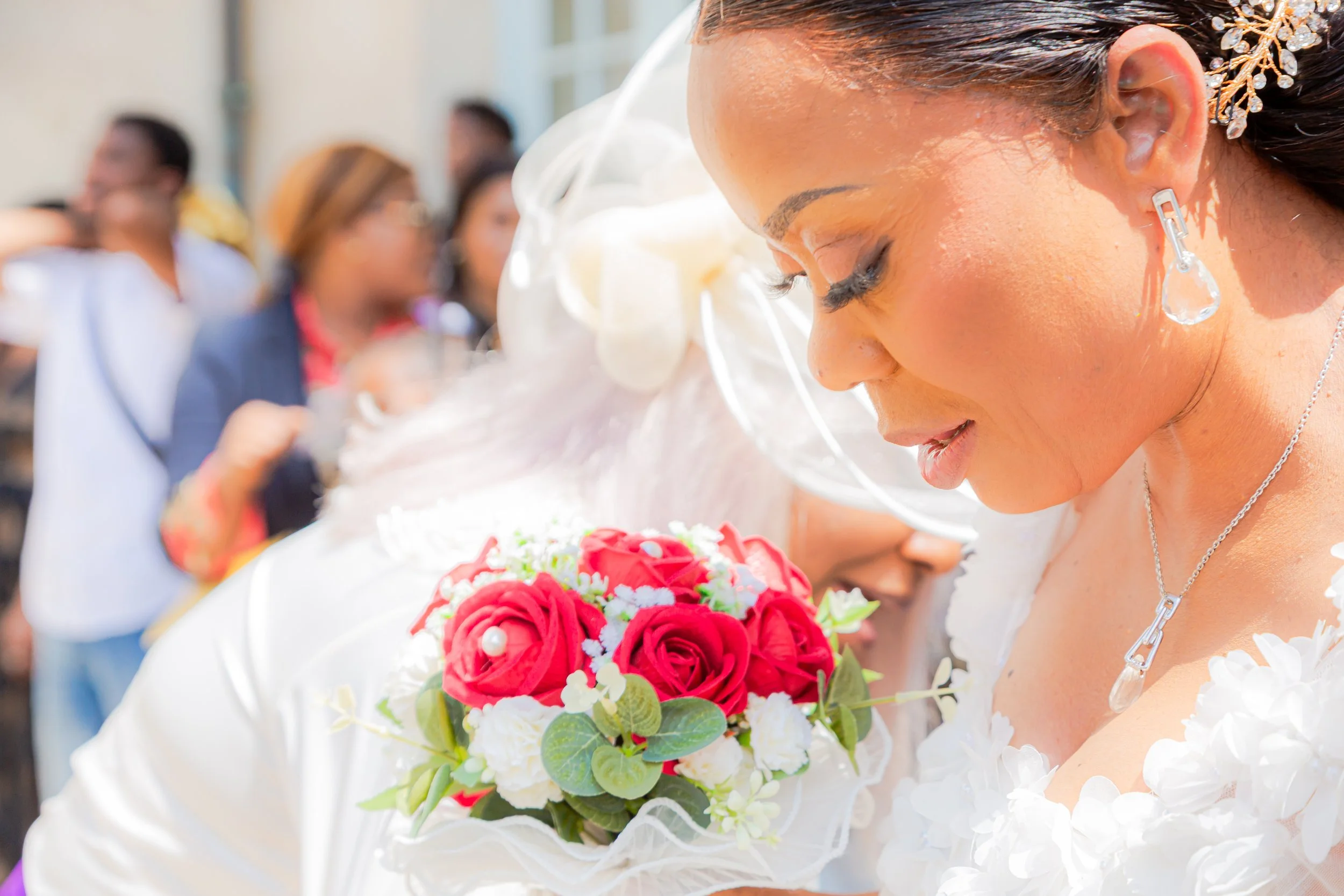 Une femme en robe blanche, avec des boucles d'oreilles en forme de goutte d'eau en cristal, tient un bouquet de fleurs rouges et blanches lors d'une cérémonie de mariage. D'autres personnes en arrière-plan regardent.