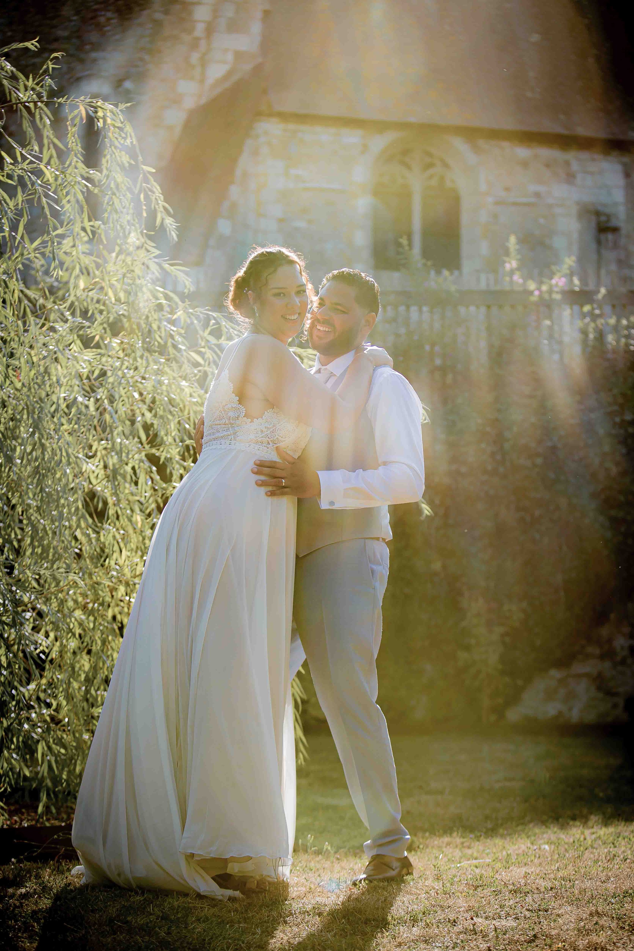 Un couple marié en habits blancs souriant dans un jardin ensoleillé devant une vieille bâtisse en pierre.