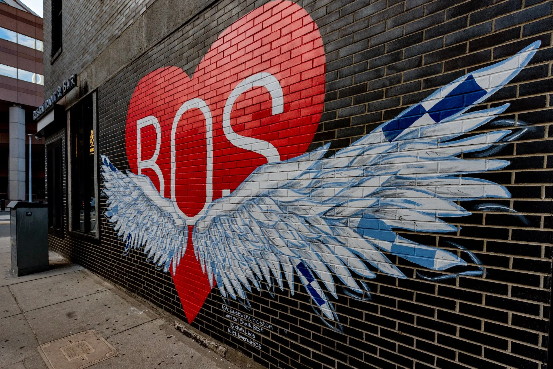 Street mural depicting a red heart with the white letters 'R', 'O', and 'S' inside it. The heart has detailed white and blue angel wings extending from its sides, painted on a dark brick wall.