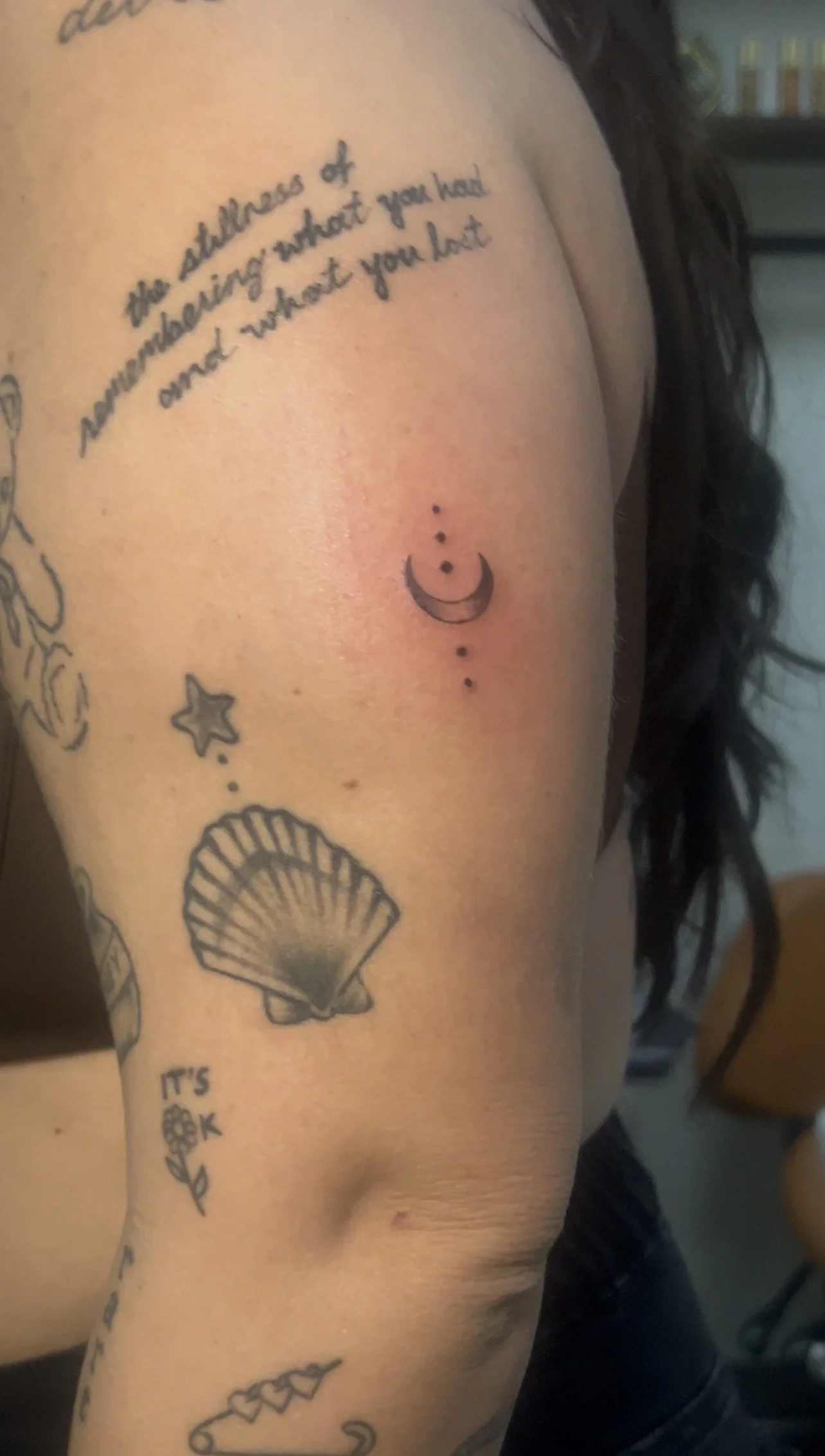 A close-up of a person's upper arm and shoulder featuring multiple tattoos, including a quote, a crescent moon with dots, a shell, stars, a flower, and other designs.