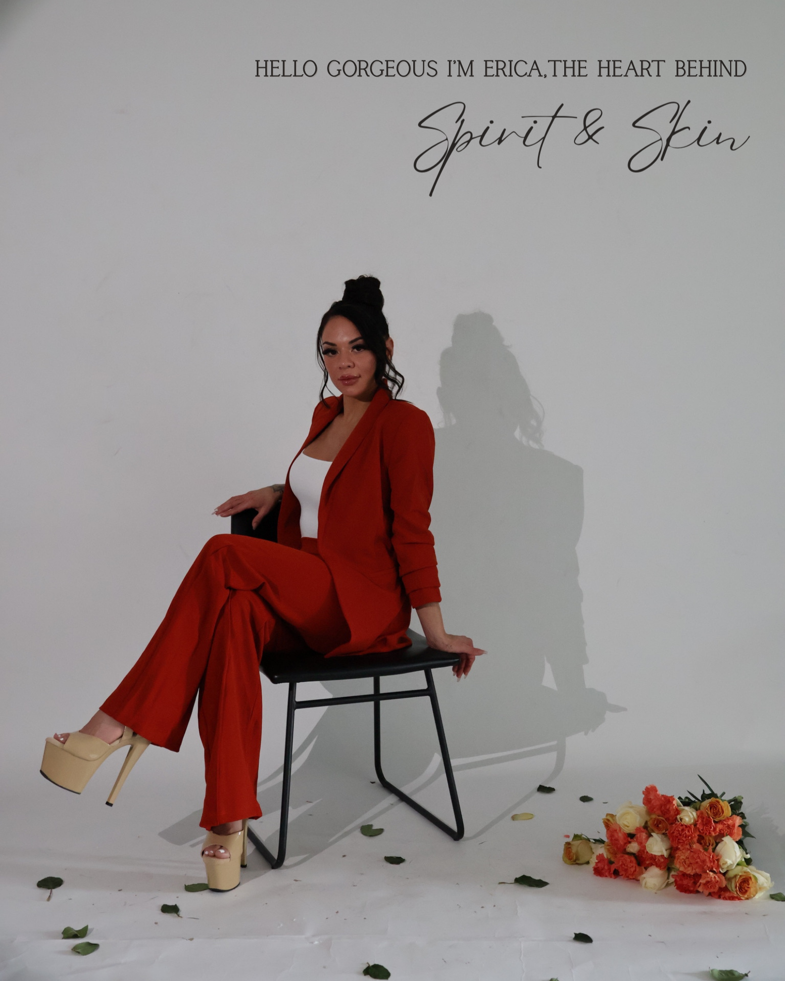A woman in a red suit with beige platform heels sitting on a black chair in a studio setting, surrounded by scattered leaves and a bouquet of roses on the floor, with a plain background and a shadow of herself on the wall. Text on the wall reads 'Hel