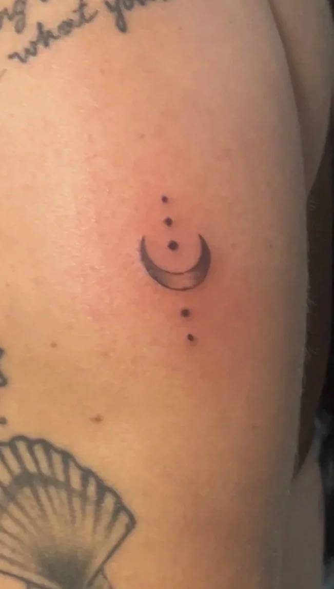 A close-up of a person's upper arm and shoulder featuring multiple tattoos, including a quote, a crescent moon with dots, a shell, stars, a flower, and other designs.