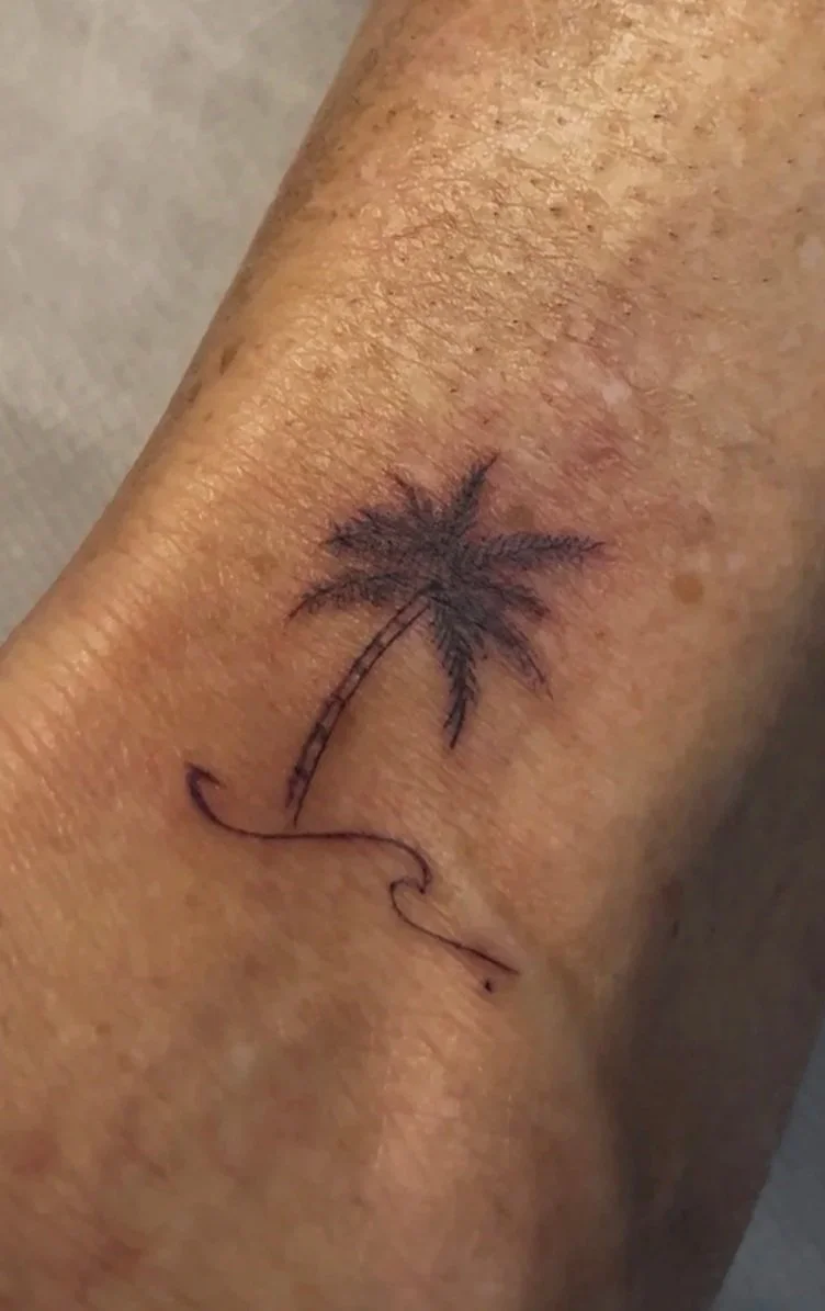 Close-up of a person's arm with a small, simple black tattoo of a palm tree on the forearm.