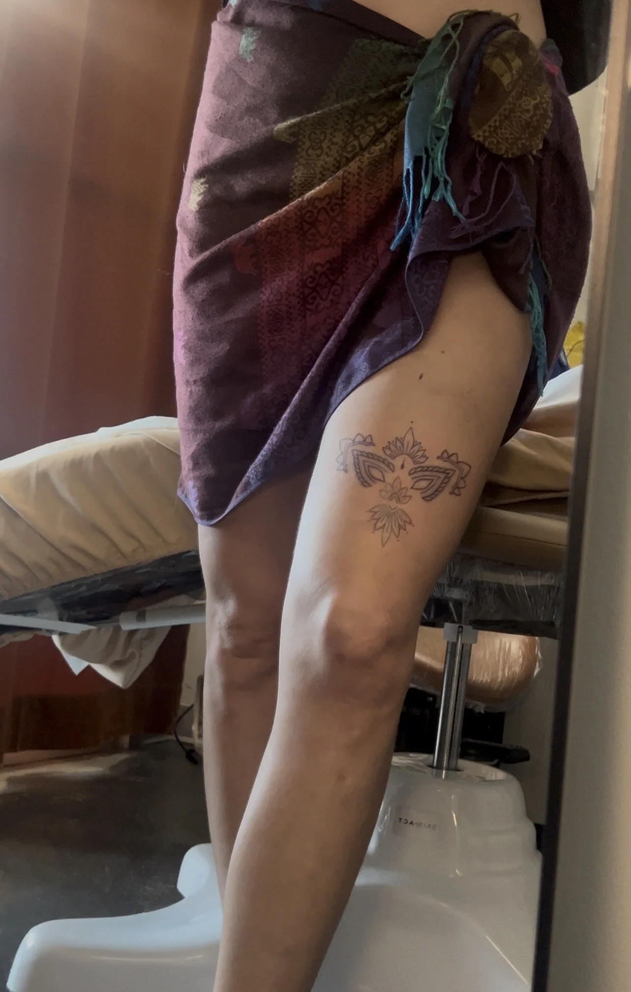A person standing in a bathroom, partially covered with a patterned cloth, showing their thigh which has a butterfly tattoo. The bathroom has a bed, a water heater, and some furniture visible.