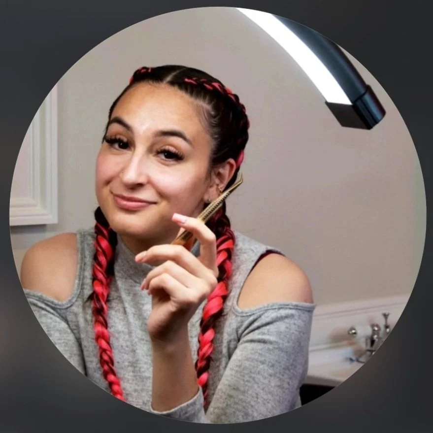 Young woman with braided hair holding a hairbrush, smiling, in front of a mirror in a bathroom.