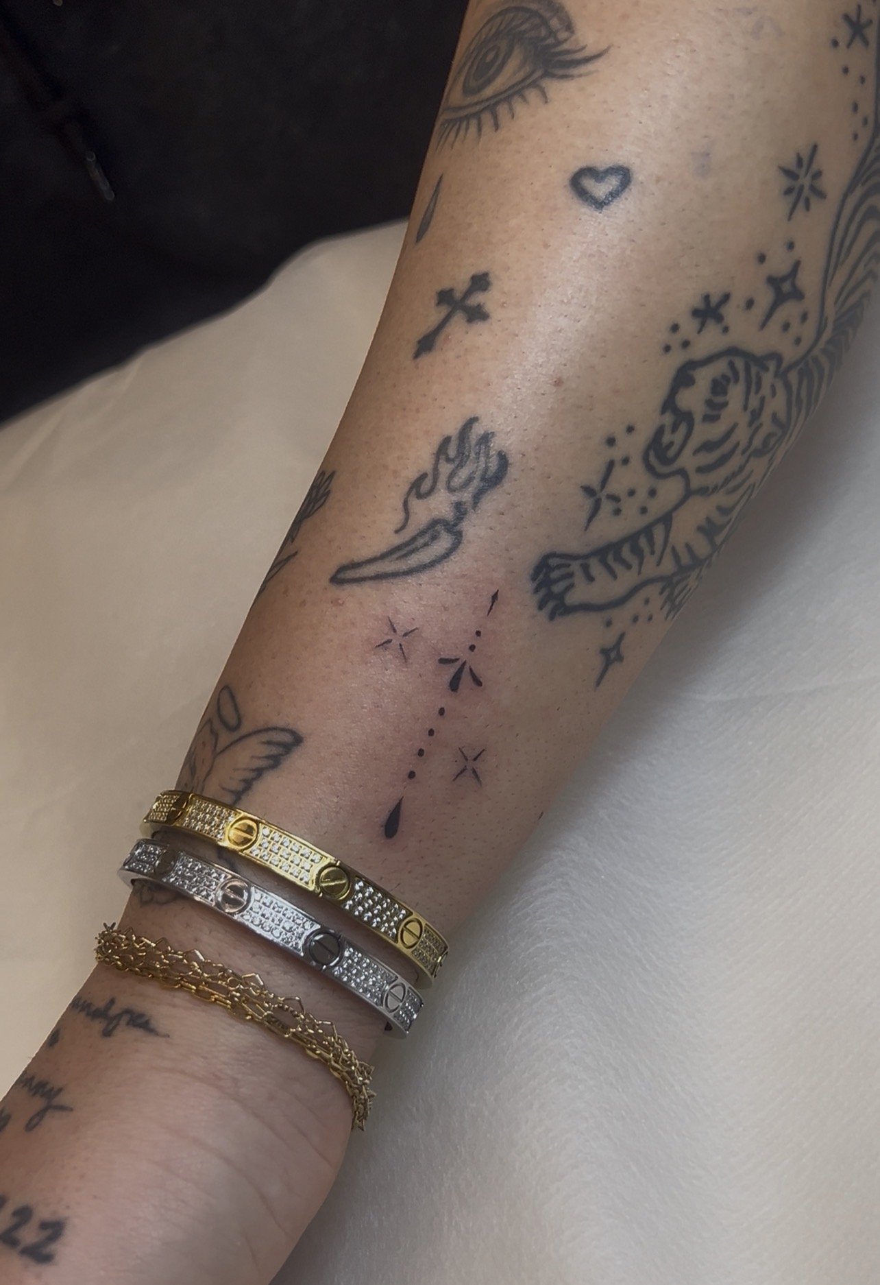 Close-up of a tattooed arm with various black ink tattoos including an eye, a heart, a cross, a flame, a tiger, and star-like designs. The arm is adorned with several bracelets: a gold chain, and three metallic slim band bracelets in gold, silver, an