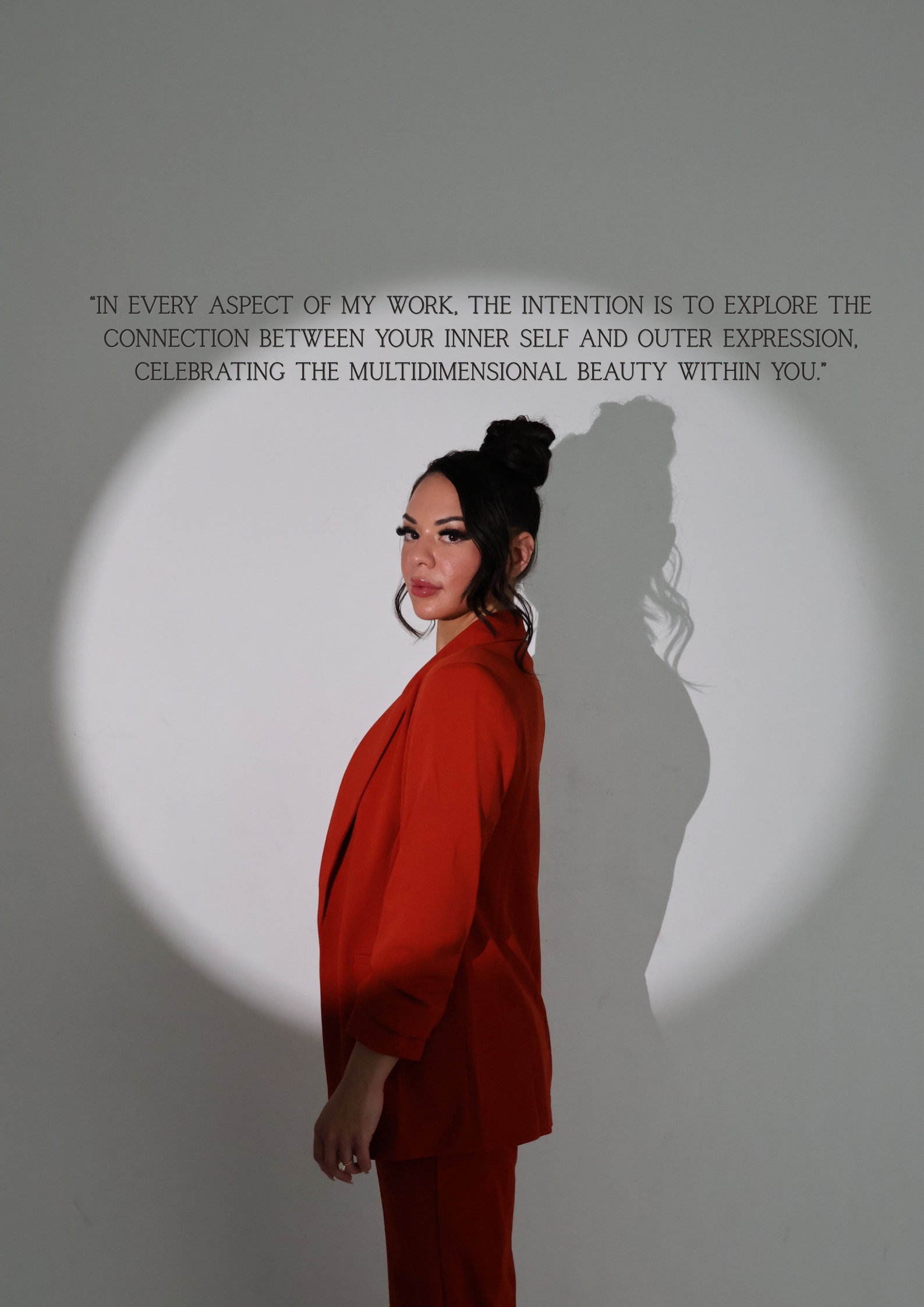 A woman with dark hair styled in a bun and long wavy hair, wearing an orange blazer and matching pants, standing against a plain background with her shadow cast on the wall behind her, and a quote on the wall above her.