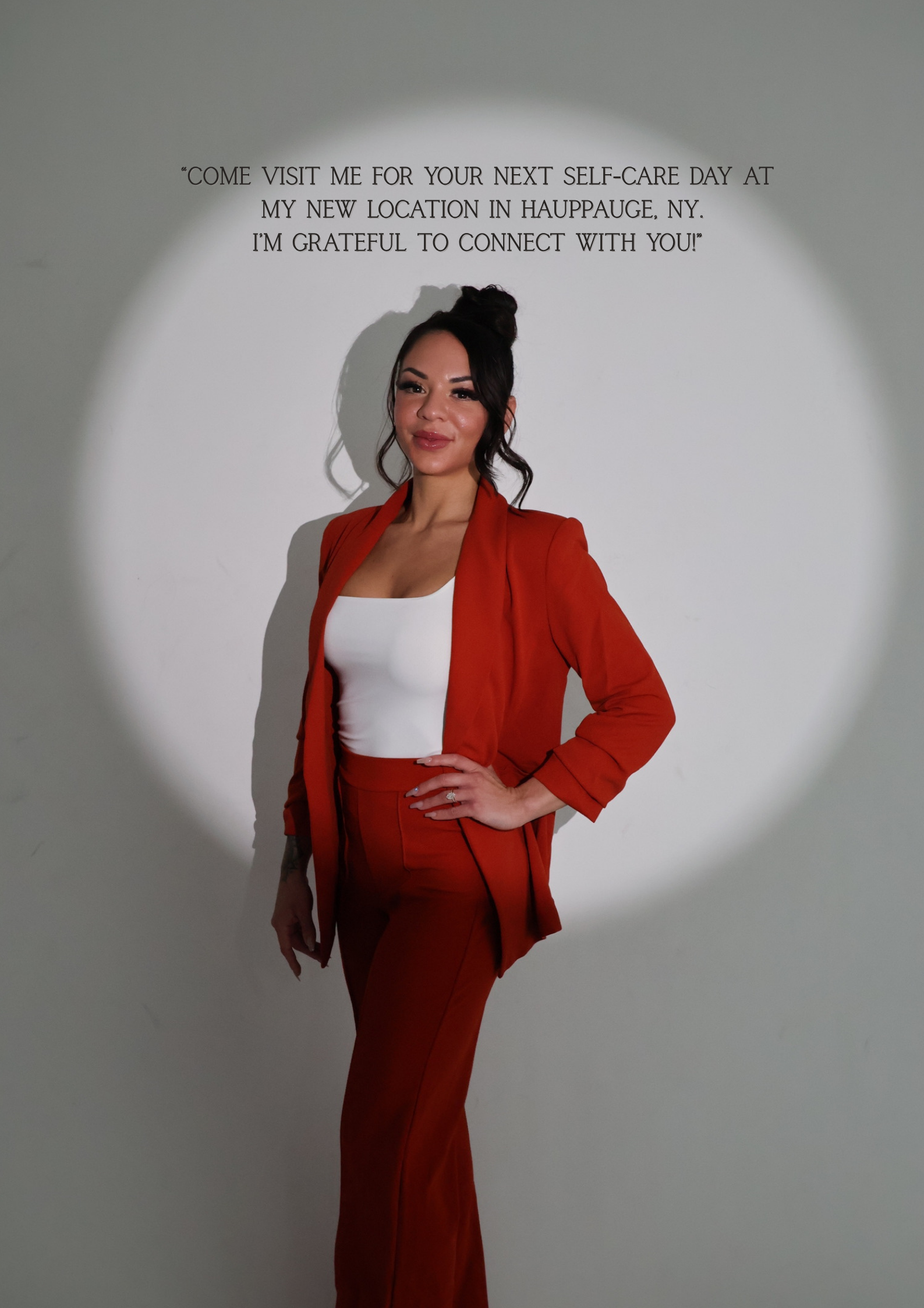 A woman with dark hair styled in a bun, wearing a red suit with matching red pants and a white top, standing in front of a white wall with black text. The text reads: "Come visit me for your next self-care day at my new location in Hauppauge, NY. I’m