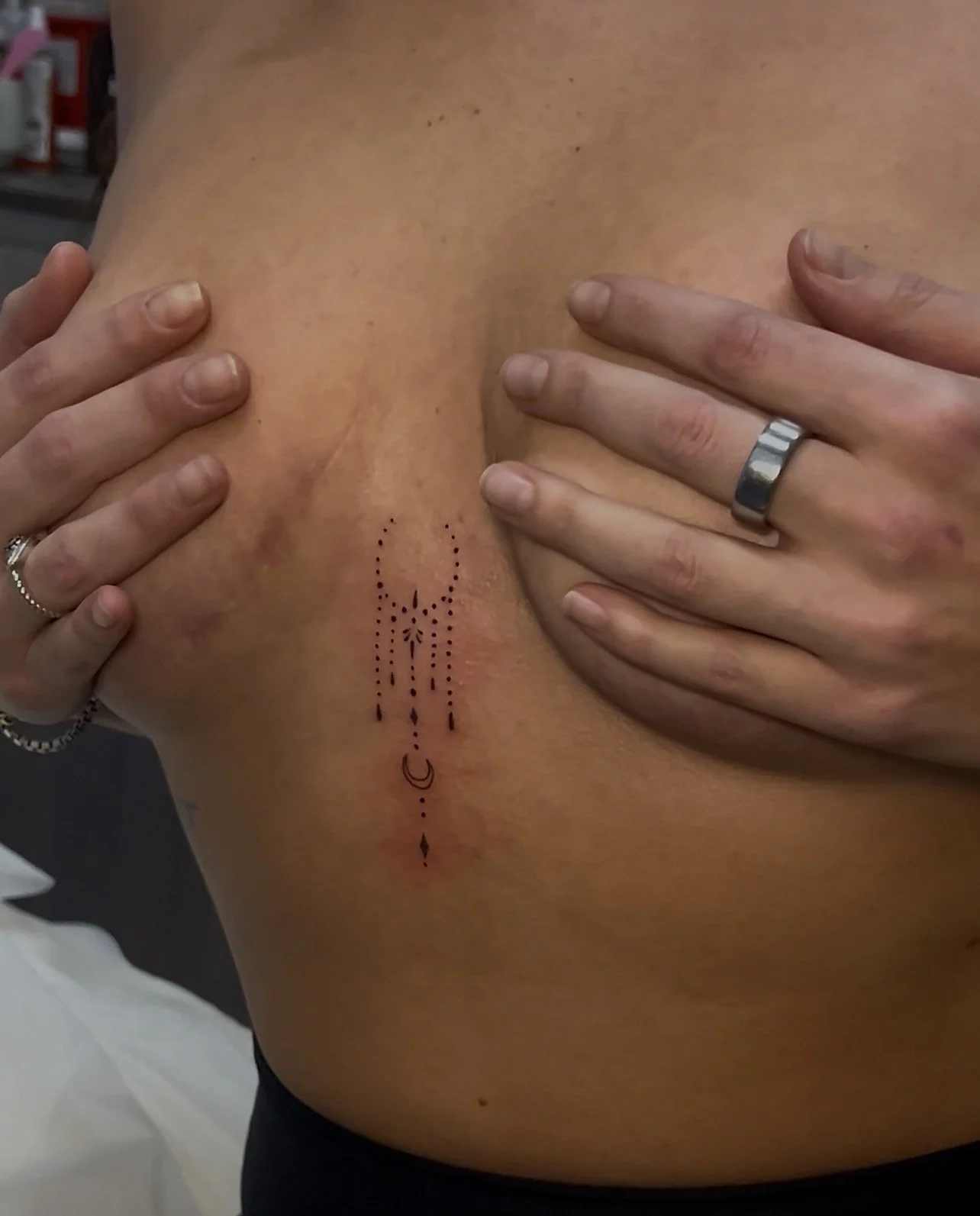 Close-up of a person with a dotted tattoo on their side, being touched by hands with rings.