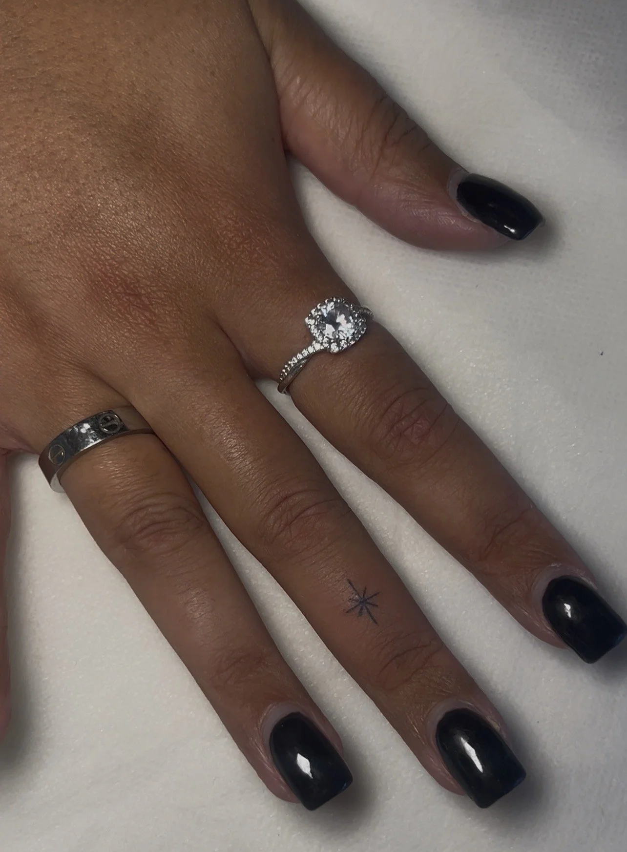 Close-up of a woman's hand with dark nail polish, wearing two rings: one on the middle finger with a large central diamond surrounded by smaller diamonds, and another on the ring finger with engraved pattern. The hand has a small star tattoo and is r