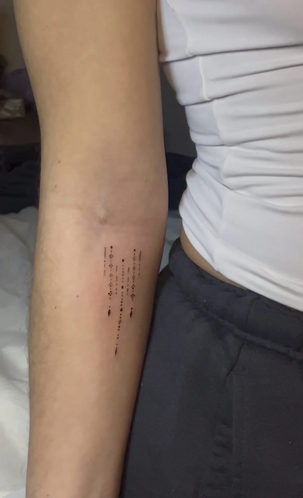 A person's arm with a tattoo of dotted lines and shapes, positioned next to them wearing a white t-shirt and dark pants, with a background of a bed and furniture.