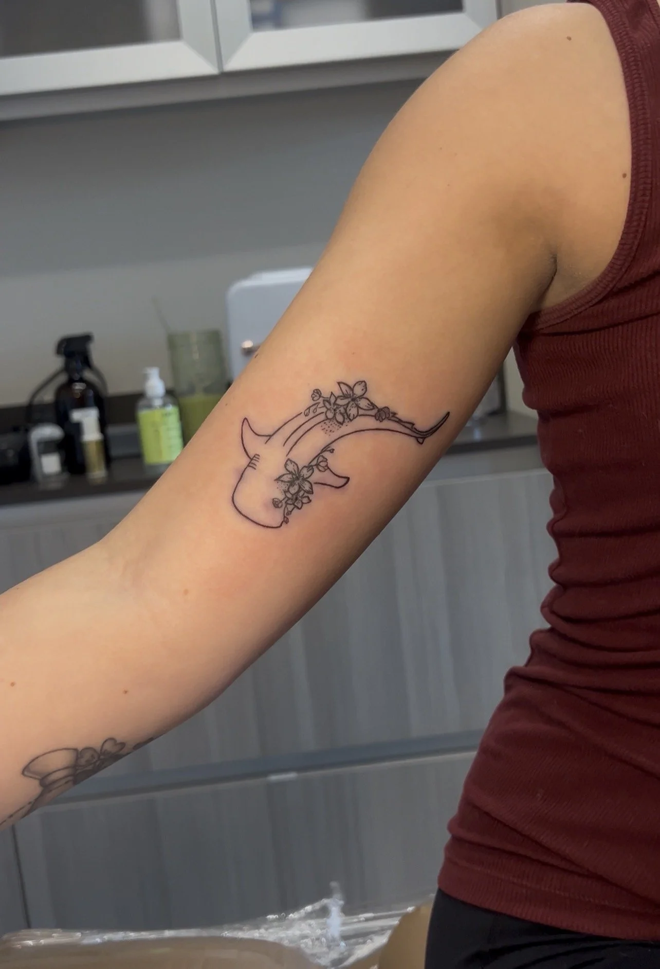A person's upper arm with a tattoo of a narwhal decorated with flowers, in a room with bathroom items visible in the background.