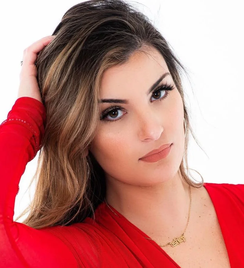 Close-up of a woman with long brown hair, blue eyes, wearing a red top, with makeup and a gold necklace, posing with her hand in her hair against a white background.