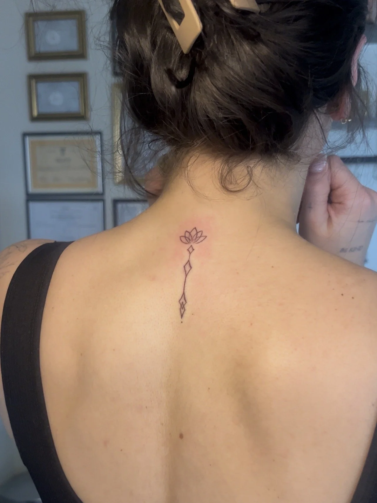 A woman with dark hair styled in a loose bun with a hair clip, showing a minimalist tattoo of a lotus flower and geometric designs on her upper back/neck area.